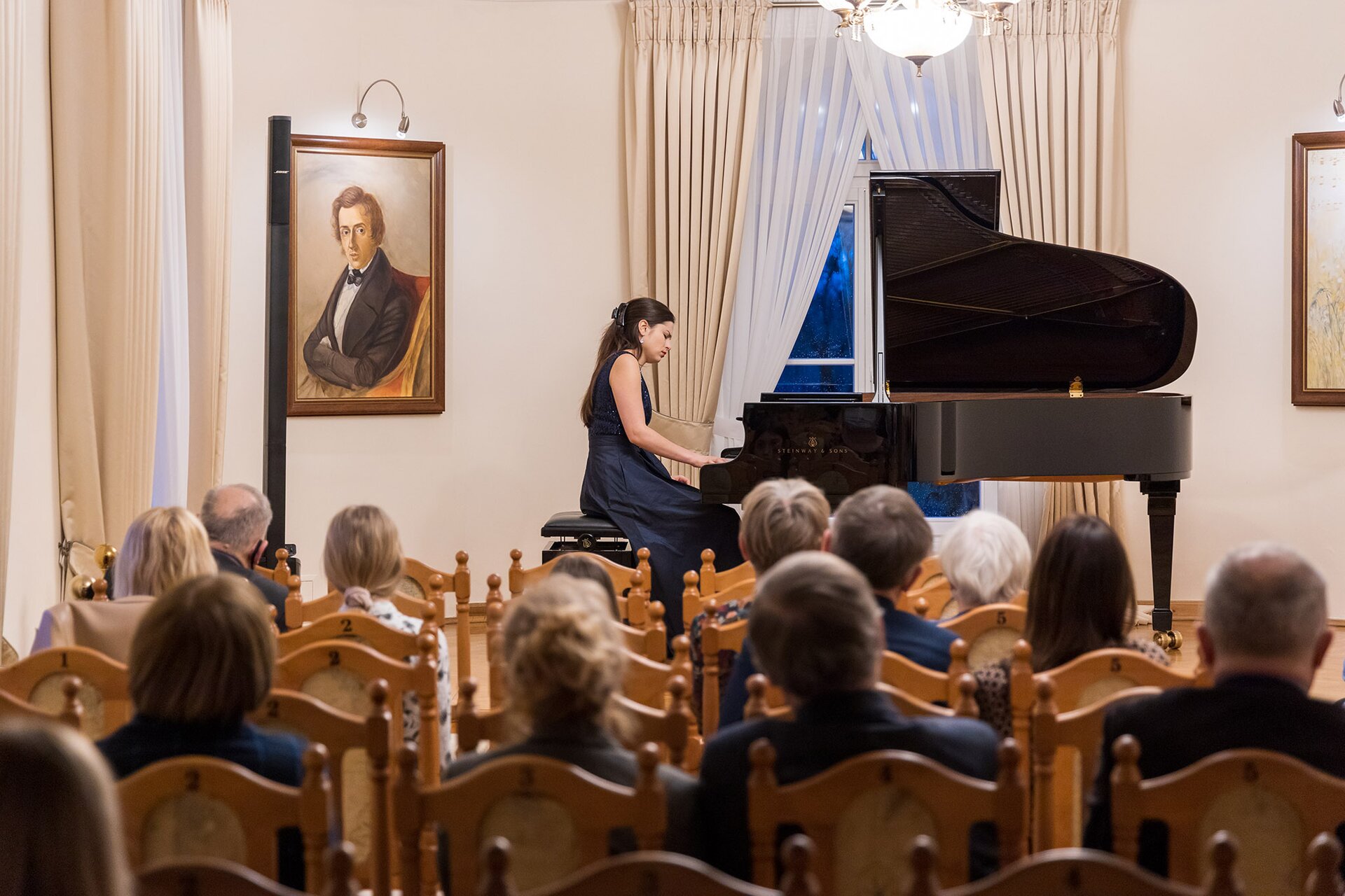 Na zdjęciu widoczna jest grupa osób siedzących na drewnianych krzesłach, zwróconych w stronę kobiety grającej na czarnym fortepianie. Ma ona na sobie długą, granatową suknię oraz upięte klamrą włosy. Osoby na zdjęciu znajdują się w pomieszczeniu o białych ścianach. Na jednej z nich wisi obraz przedstawiający elegancko ubranego mężczyznę. Okna są ozdobione białymi i beżowymi firanami.