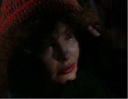 Zdjęcie ukazuje twarz kobiety w średnim wieku. Czoło przysłonięte jest włosami, oczy kobiety są także częściowo przesłonięte włosami. Włosy są bujne, kręcone, długie i ciemne. Kobieta ma szeroki nos, duże, pełne usta. Na głowie ma założoną wełnianą czapkę.