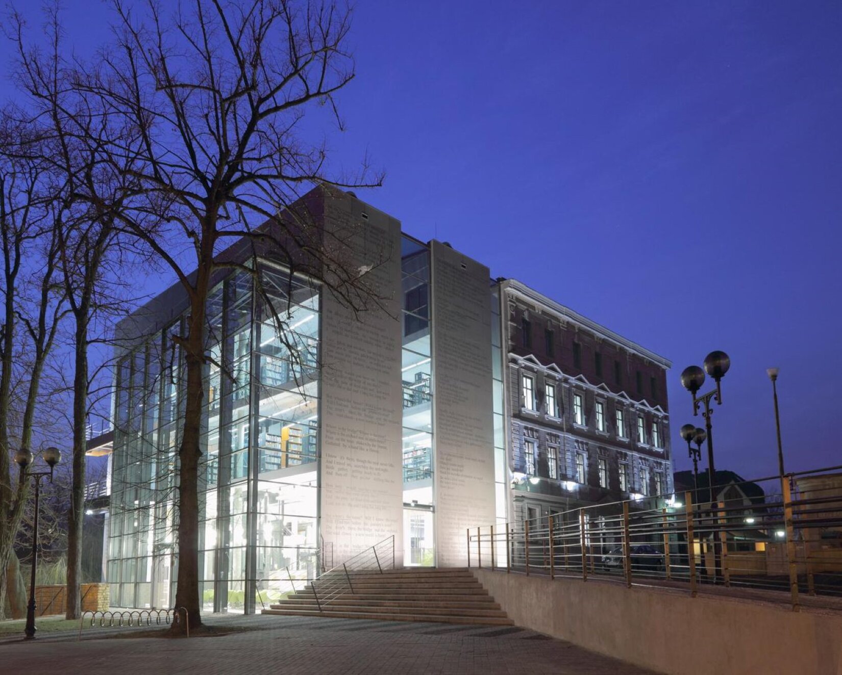 Prostokątna fotografia ukazuje duży budynek składający się z dwóch części. Pierwsza z nich jest nowoczesna. Jej ściany są szare oraz posiadają napisy. Większość ścian jest przeszklona. W pomieszczeniach budynku ustawione zostały regały zapełnione książkami. Druga część budynku jest szara, ozdobiona łukami, fasadami oraz ornamentami. Jej ściany podświetlone są punktowymi światłami. Do budynku prowadzą schody oraz podjazd otoczone srebrnymi barierkami. Wokół budynku ustawione są latarnie. Z lewej strony fotografii zamocowany jest stojak na rowery. Przy nim zasadzone są drzewa. 