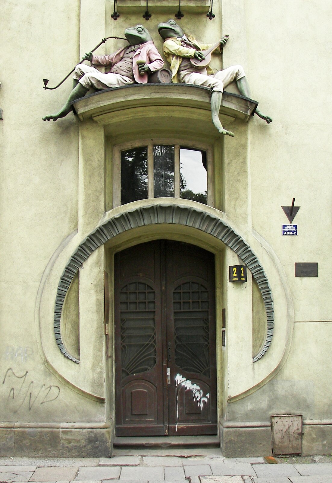 Zdjęcie przedstawia wysokie drzwi, wokół których umieszczono półokrągłe zdobienie w zielonym kolorze. Nad zdobieniem znajduje się okno, a nad oknem - wystający daszek. Na nim dwie rzeźby siedzących żab. Żaba po lewej stronie jest ubrana w różową marynarkę, brązową koszulę i spodnie za kolano. Trzyma w ręce kielich. W drugiej ręce trzyma fajkę. Żaba po prawej stronie gra na mandolinie. Ma na sobie żółtą marynarkę, brązową koszulę i spodnie za kolano.