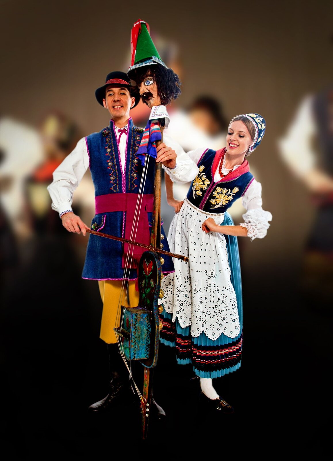 Fotografia przedstawia prezentacje tradycyjnego stroju kaszubskiego. Na fotografii znajduje się kobieta oraz mężczyzna. Kobieta ubrana jest w czarne buty, długą niebieską suknię z białym fartuszkiem, białą koszulę oraz chustę na głowie. Mężczyzna ubrany jest w czarne kozaki, luźne żółte spodnie, niebieską marynarkę oraz białą koszulę. Na głowie mężczyzny znajduje się kapelusz. Mężczyzna w dłoniach trzyma tradycyjny kaszubski instrument nazywany diabelskimi skrzypcami. Instrument wyglądem przypomina duże skrzypce, jednak na jego czubku znajduje się głowa kukły. Tło obrazka jest ciemne oraz rozmazane.