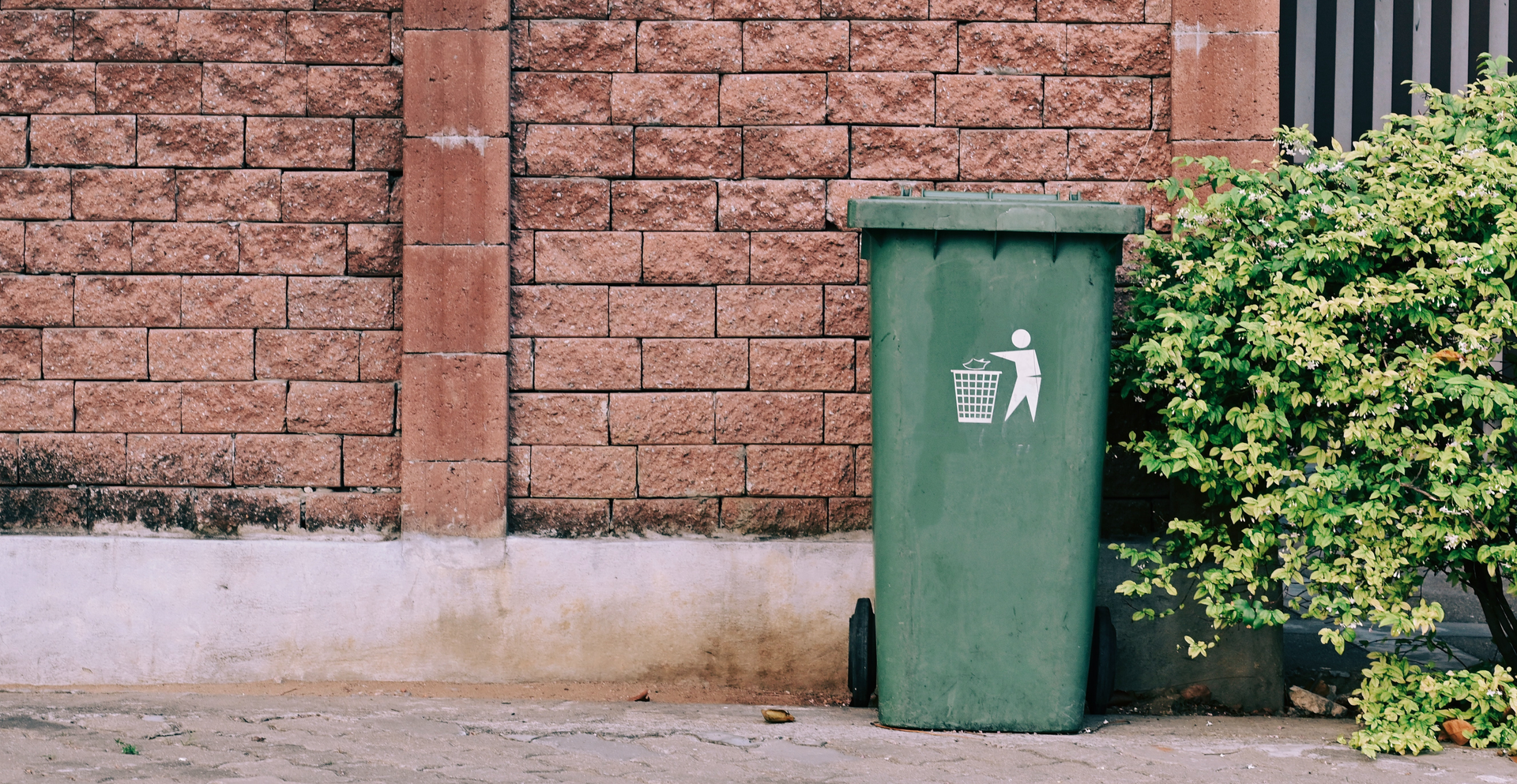 Zdjęcie przedstawia kosz na śmieci stojący przy ścianie budynku.