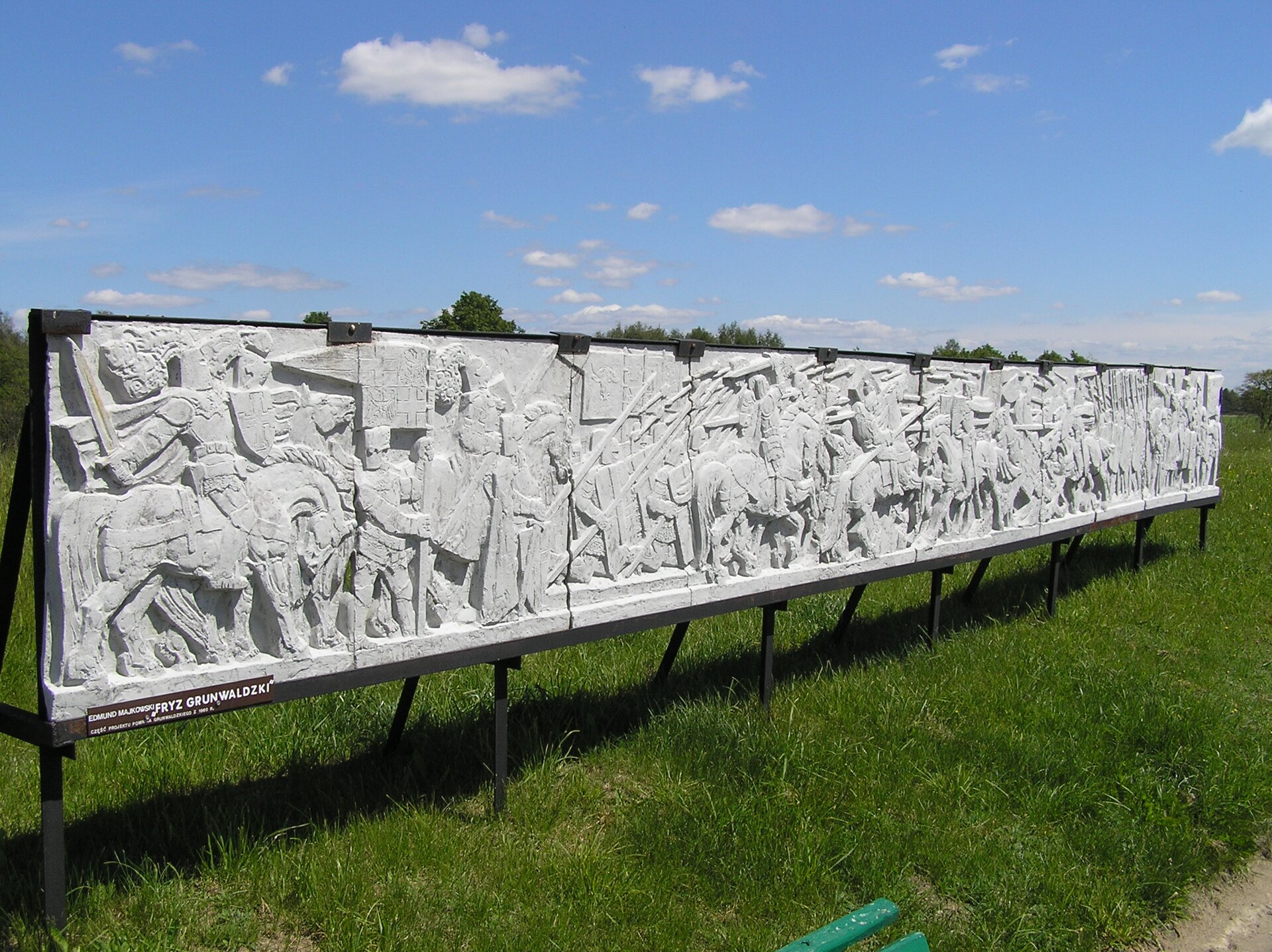 Na zdjęciu przedstawiono stojące na trawie kamienne tablice, na których wyryto w formie płaskorzeźb sceny z bitwy pod Grunwaldem. Ukazują one uzbrojonych rycerzy na koniach podczas walk. W głębi widać niebieskie niebo.