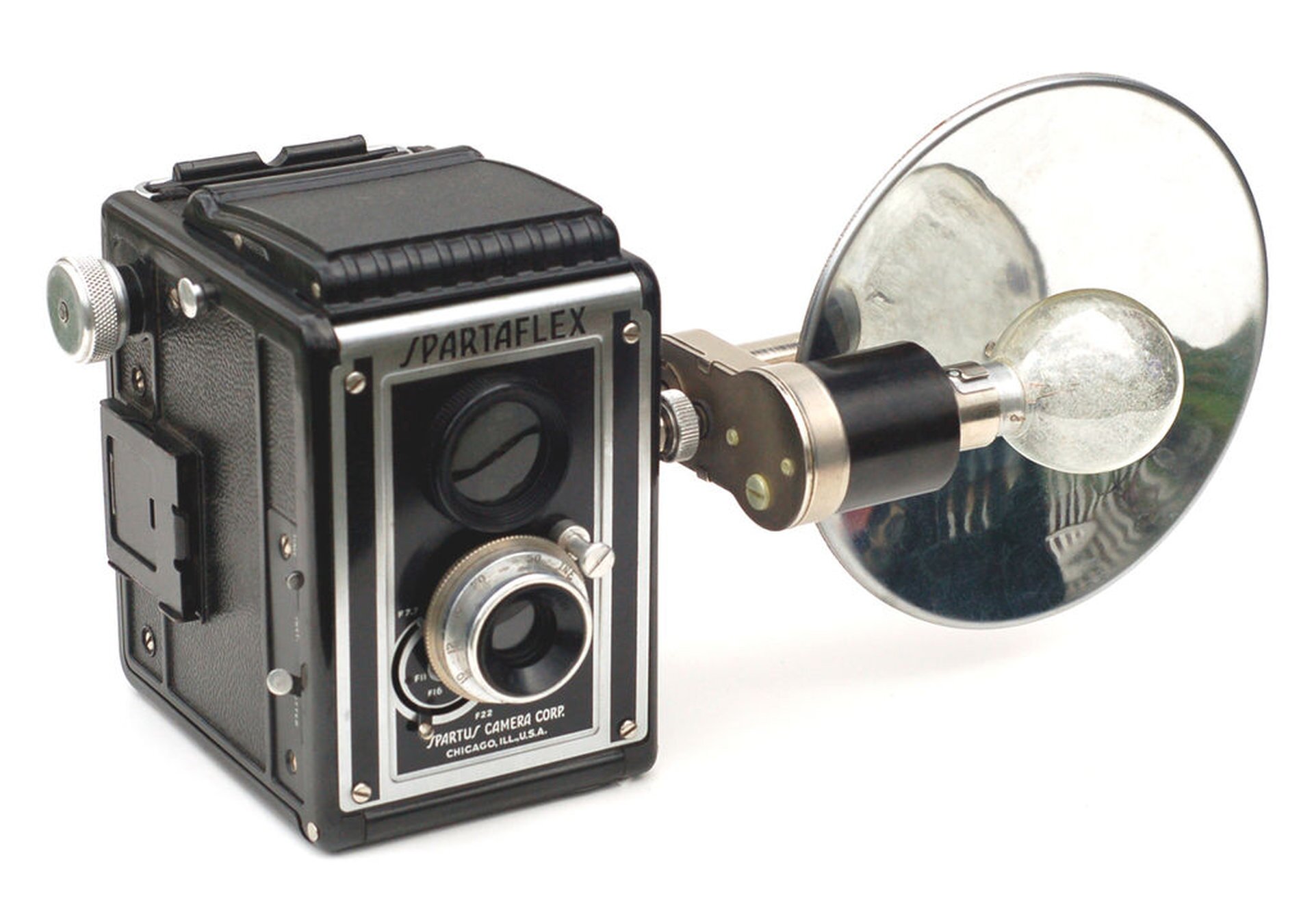 Rys. a. Na zdjęciu przedstawiono aparat fotograficzny pochodzący z lat czterdziestych dwudziestego wieku. Korpus aparatu ma kształt prostopadłościennego pudełka. Na jednej ścianie znajdują się dwa obiektywy w kształcie walca. Na sąsiadującej ścianie zamontowane jest ramię, do którego podłączony jest flesz. Jest to żarówka, za którą znajduje się okrągłe lustro, mające za zadanie odbić całe światło od flesza.