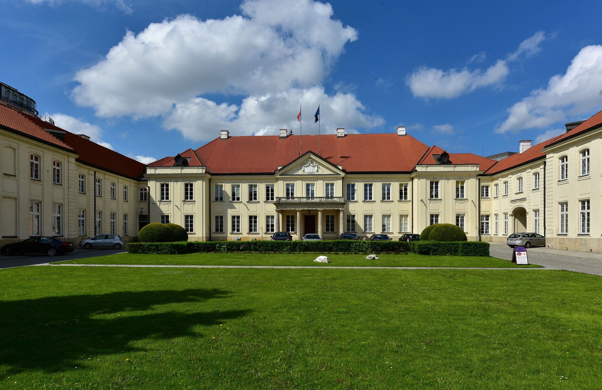 Zdjęcie przedstawia niewysoki, zabytkowy, dwukondygnacyjny budynek w stylu klasycystycznym. Budynek jest w kształcie litery U: jest tu część główna oraz dwa boczne skrzydła. Do części głównej prowadzi wejście poprzez portyk z kolumnadą (4 kolumny). Na portyku wspiera się niewielki taras wykończony betonową barierką oraz ozdobnym frezem poniżej. Okna w budynku są wysokie i wąskie. W oknach są szprosy. Nad trzema oknami - wejściami z tarasu do budynku znajduje się tympanon, w którym umieszczony jest kamienny herb rodziny Potockich. Nad tympanonem znajdują się dwa maszty, na których powiewają flagi Polski i UE. Wejście z tarasu do budynku jest nieco wysunięte do przodu. Część wieńcząca główny budynek z prawej i lewej strony również jest nieco wysunięta do przodu. Dach jest spadzisty, pokryty dachówką ceramiczną w rudym kolorze. Przed budynkiem na pierwszym planie znajduje się zielony trawnik. 
