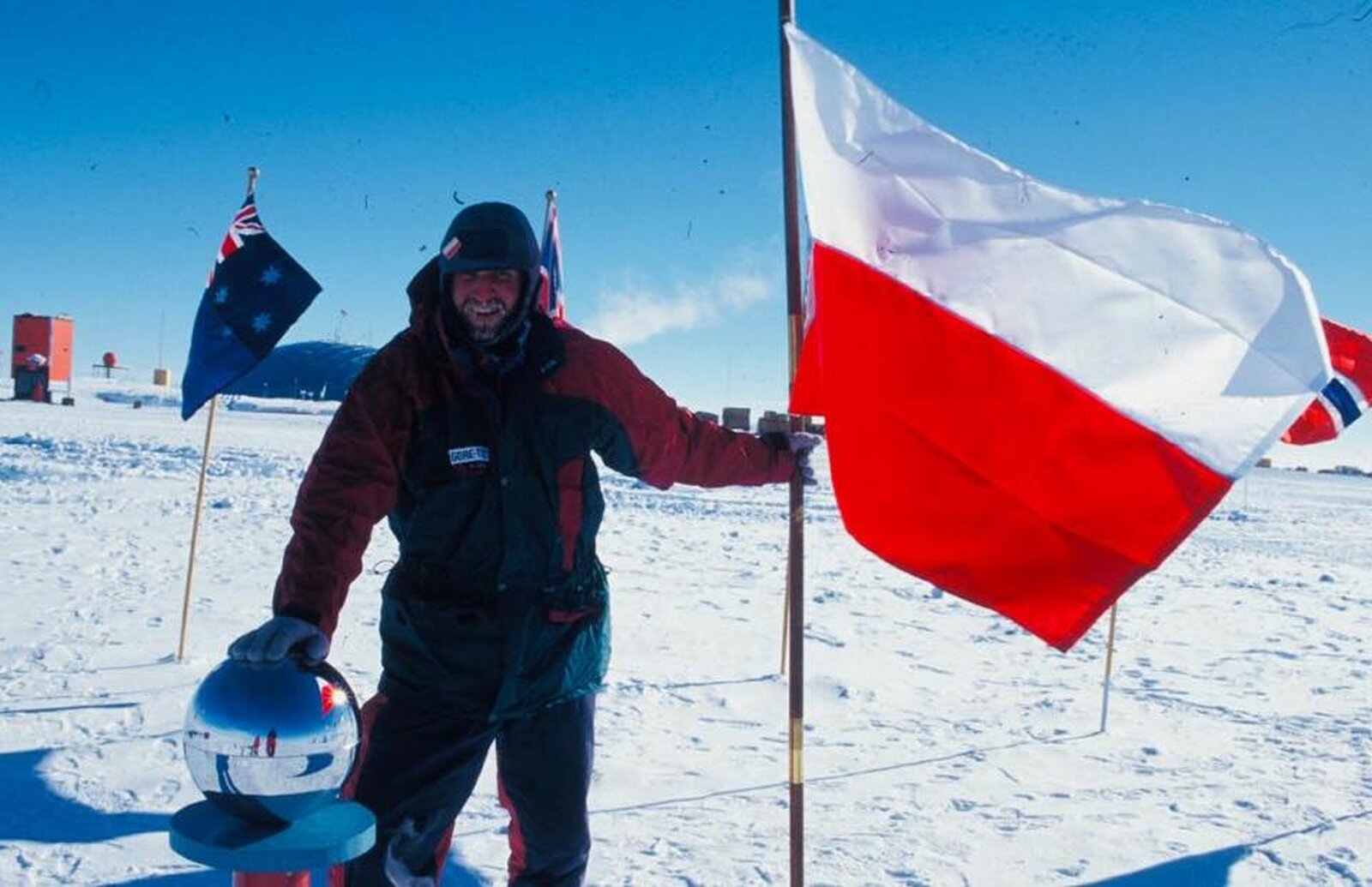 Fotografia przedstawia mężczyznę ubranego w zimowy kombinezon z czapką. W lewej ręce trzyma flagę Polski wbitą w ziemię. Prawą ręką opiera się o szklaną kulę. Dookoła śnieg. w tle inne flagi. 