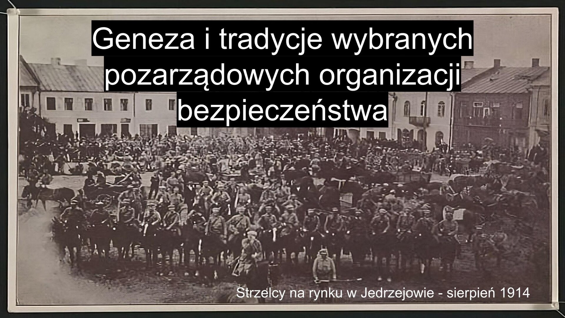 Slajd. Na środku czarno‑białe zdjęcie dużą grupę młodych mężczyzn w mundurach żołnierskich, którzy stoją na placu otoczonym przez piętrowe budynki. W lewym dolnym rogu biały podpis: „Strzelcy na rynku w Jędrzejowie - sierpień 1914”. U góry slajdu nagłówek: „Geneza i tradycje wybranych pozarządowych organizacji bezpieczeństwa”.