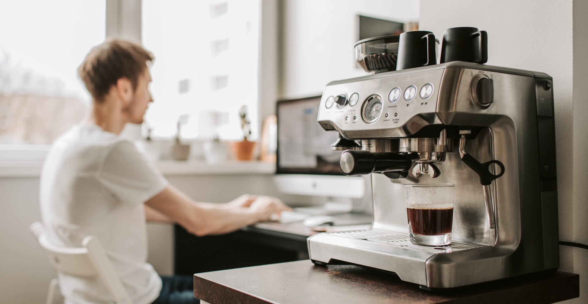 Zdjęcie przedstawia ekspres do kawy. W tle widoczny jest mężczyzna siedzący przed komputerem.