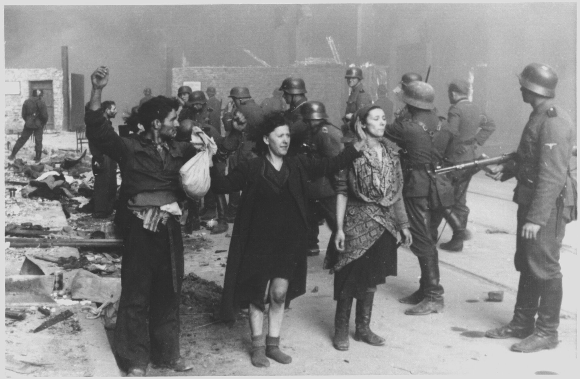 Czarno‑biała fotografia przedstawia grupę członków żydowskiego ruchu oporu aresztowanych przez niemieckich żołnierzy. żydzi mają podniesione do grupy ręce i są otoczeni przez niemieckich żołnierzy uzbrojonych w karabiny maszynowe. W oddali znajdują się zniszczone i płonące kamienice. 