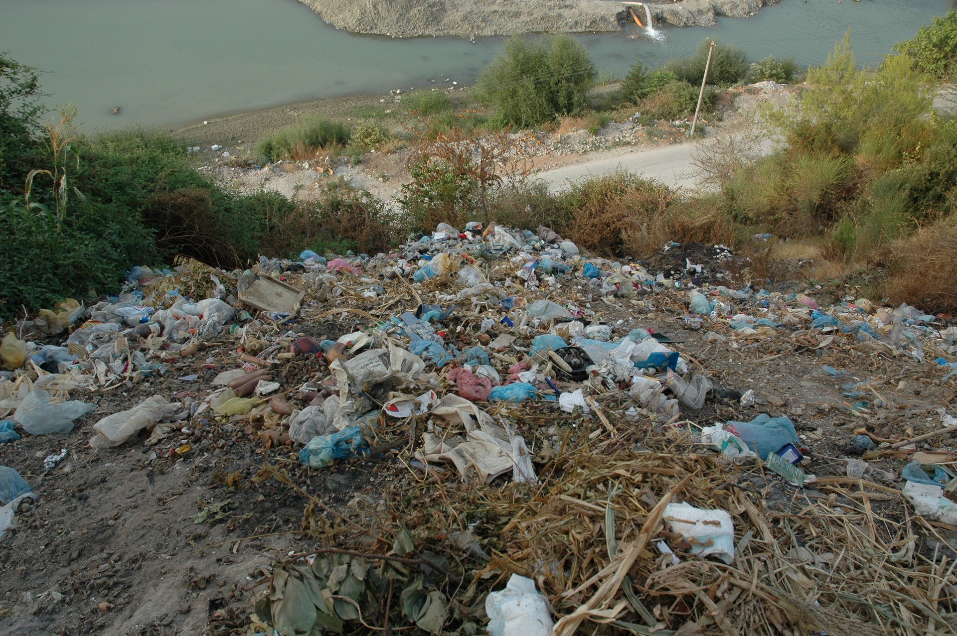 Fotografia prezentuje dzikie wysypisko. Widoczne porozrzucane kolorowe śmieci na górce. Poniżej górki widoczna droga oraz woda.