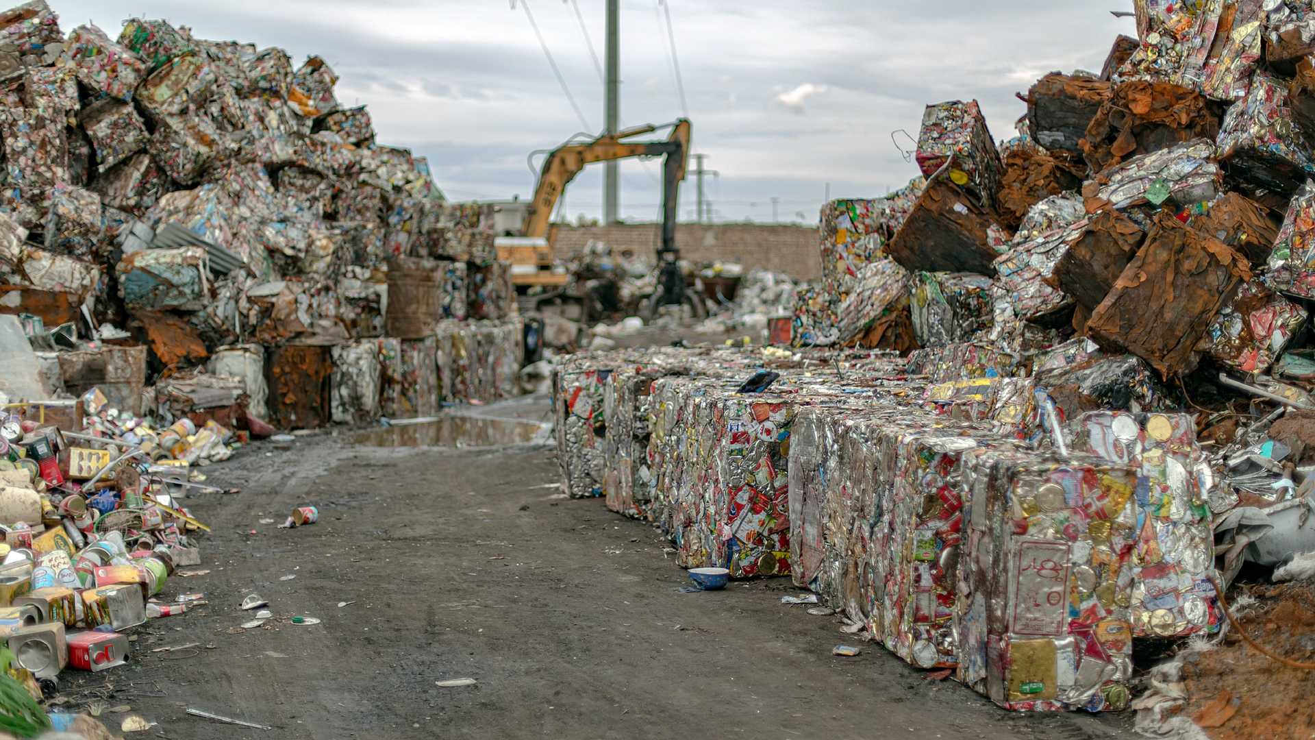 Zdjęcie przedstawia góry odpadów. Widoczne są paczki sprasowanych odpadów. W tle widoczny jest wysięgnik koparki.