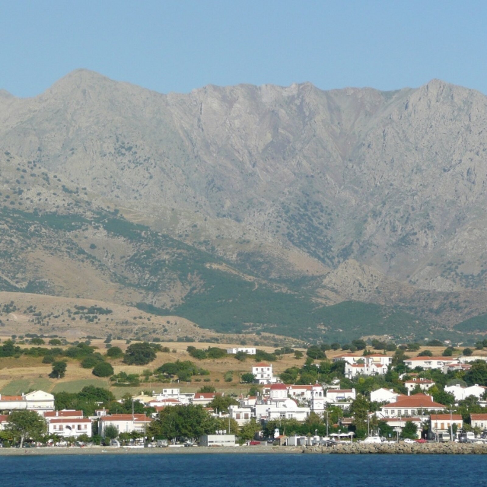 Ilustracja przedstawia kolorową fotografią greckiego miasteczka Chora Samothrakis. Na pierwszym planie ukazana jest błękitna tafla wody. Za nią znajdują się zabudowania. Białe budynki z czerwonymi dachami usytuowane są wśród zieleni. Za miastem rozpościera się teren równinny porośnięty gdzieniegdzie zaroślami. Tło stanowi skalna ściana wysokich gór.