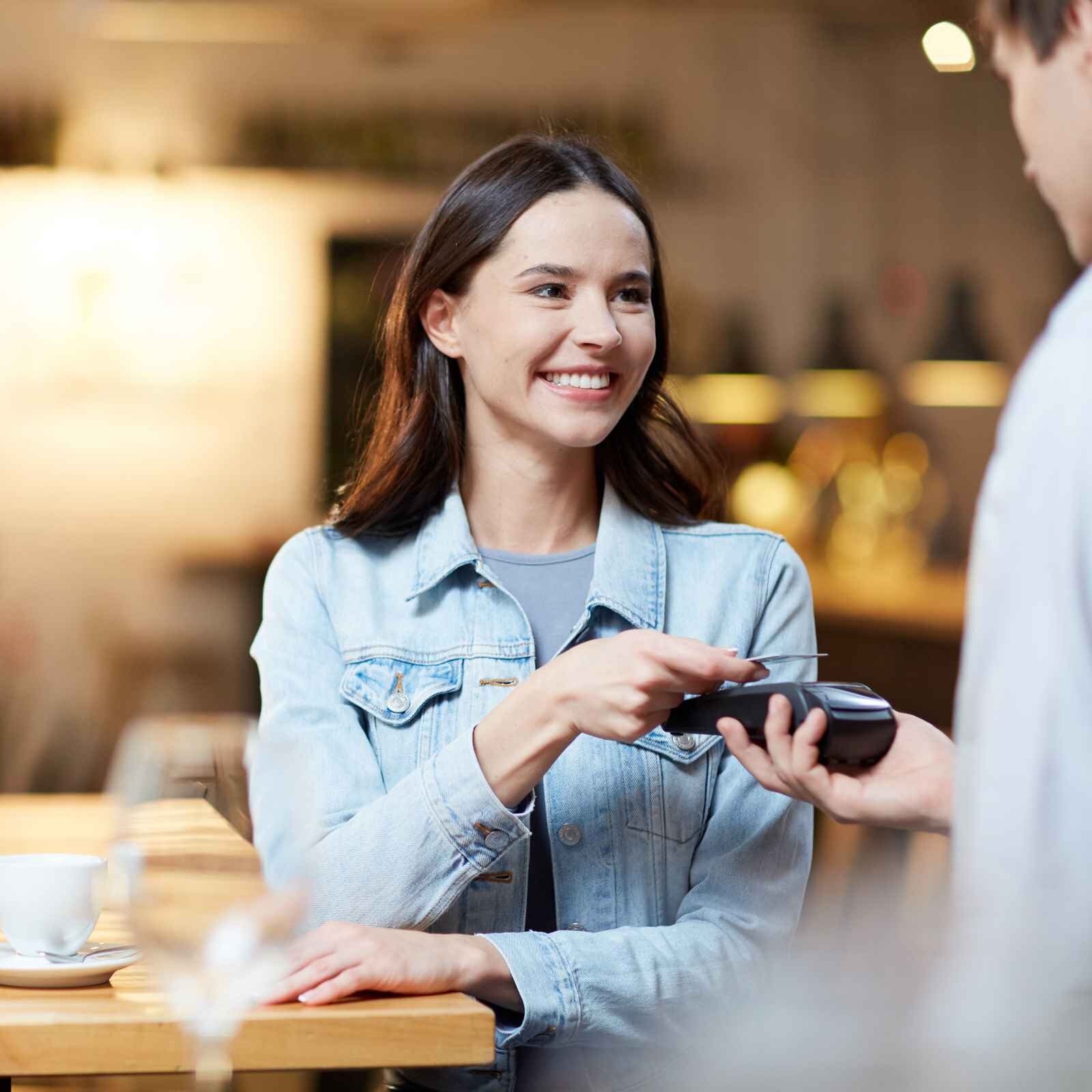 Zdjęcie przedstawia kobietę siedzącą przy stoliku w restauracji. Naprzeciw niej stoi mężczyzna z terminalem płatniczym. Kobieta ma w ręku kartę i trzyma ją nad terminalem.