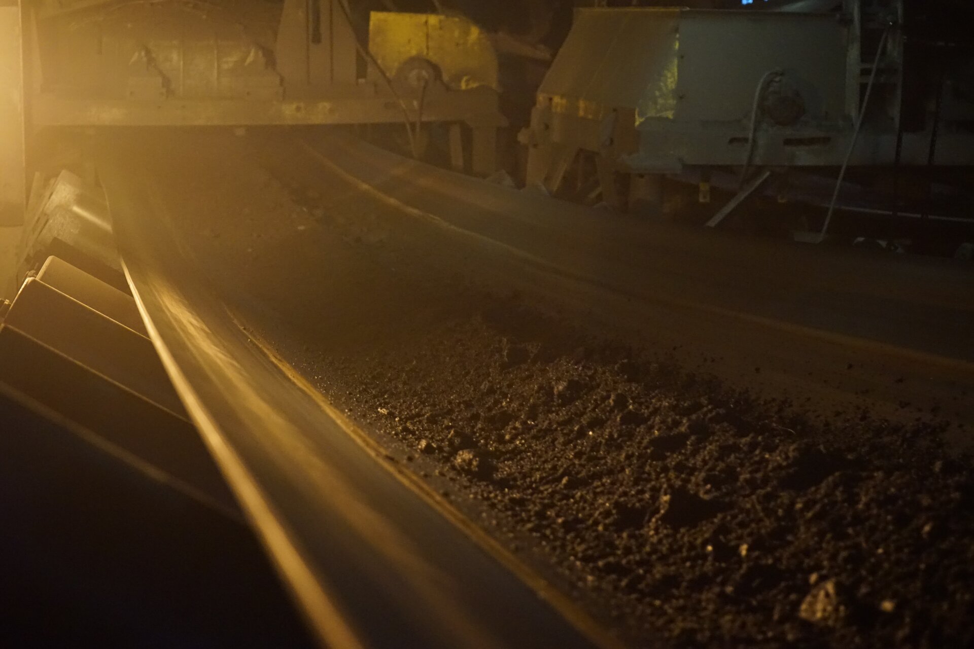 Zdjęcie przedstawia taśmę przenośnika. Transportowane są nią rozdrobnione ziarna węgla. Nad taśmą unosi się pyl węglowy. Poniżej zdjęcia znajduje się przycisk multimedialny z nagraniem audio. Treść nagrania jest zgodna z tytułem zdjęcia.