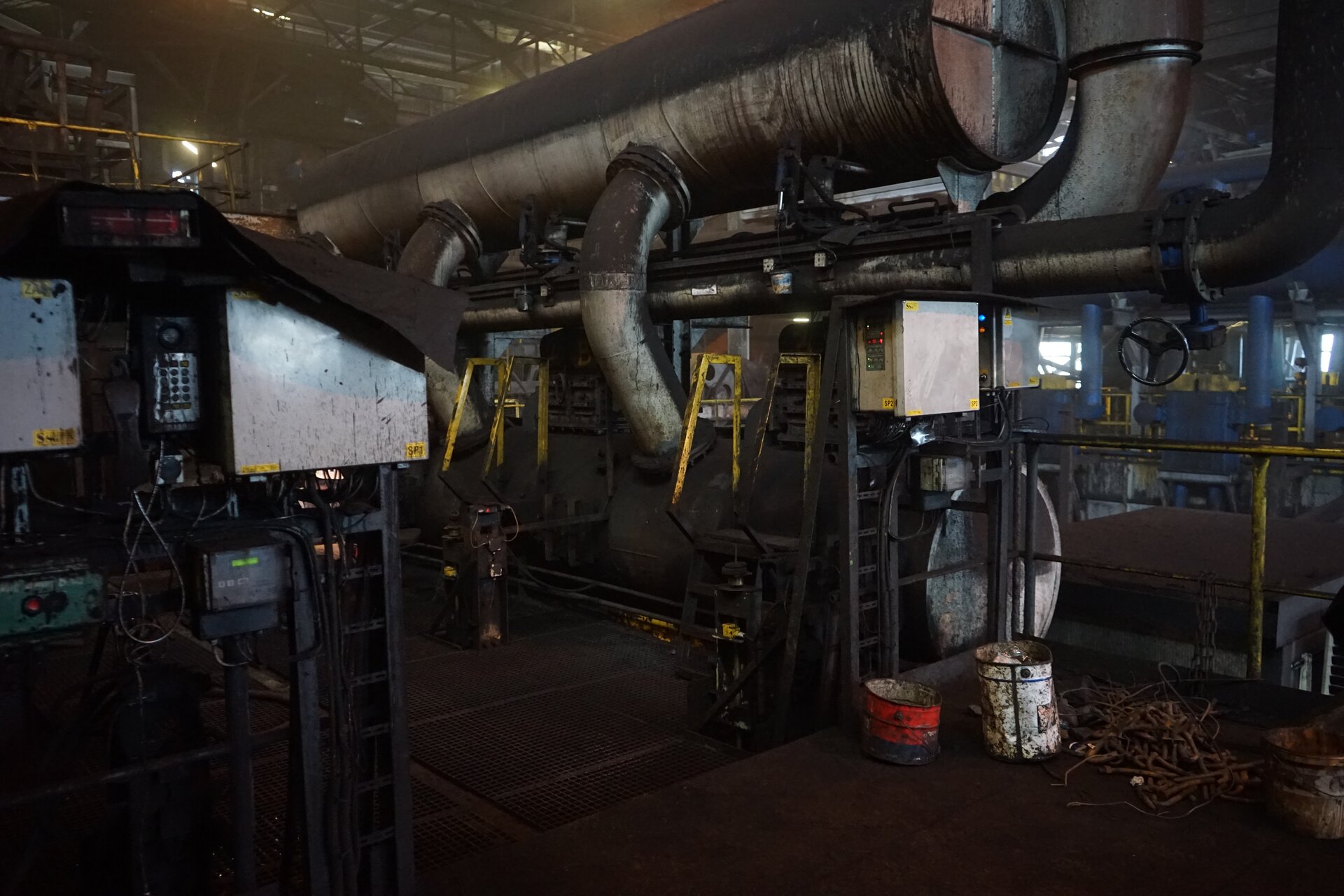 Na zdjęciu widoczna jest osadzarka widziana z boku. Osadzarka służy do wzbogacania głównie surowego węgla kamiennego oraz pozyskiwania żwiru i piasku przy równoczesnym wydzielaniu zanieczyszczeń organicznych i mineralnych. Centralnym elementem osadzarki jest podłużny cylindryczny zbiornik. Zbiornik jest o kształcie stalowej rury o dużej średnicy. Z boku zbiornika odchodzą dwie rury. Tuty są połączone ze zbiornikiem połączeniem kołnierzowym. Na dole rur znajdują się półokrągłe zbiorniki. Pomiędzy nimi znajdują się żółte stalowe pręty. Pręty są o kształcie trapezu. Przed osadzarką stoi urządzenie o przekroju trapezu równoramiennego.Osadzarka znajduje się w dużej hali przemysłowej.