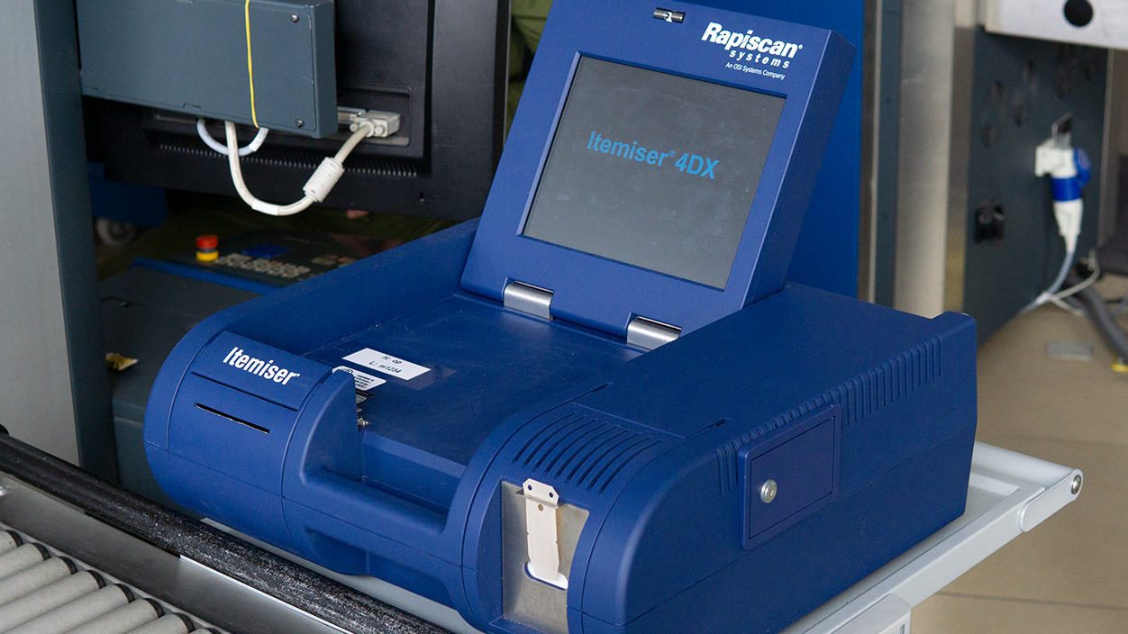 Zdjęcie przedstawia E Te De. W nawiasie ekspolsif trejs detekszyn. czyli urządzenie do wykrywania śladowych ilości materiałów wybuchowych. Jest to duża niebieska maszyna, z której wystaje ekran. Przed ekranem w urządzeniu jest wgłębienie na bagaż. Na ekranie jest napis. Itemajser cztery de iks. Nad ekranem, na rmace jest napis. Rapiskan. W lewym dolnym rogu urządzenia, poza wgłębieniem jest napis. Itemajser.