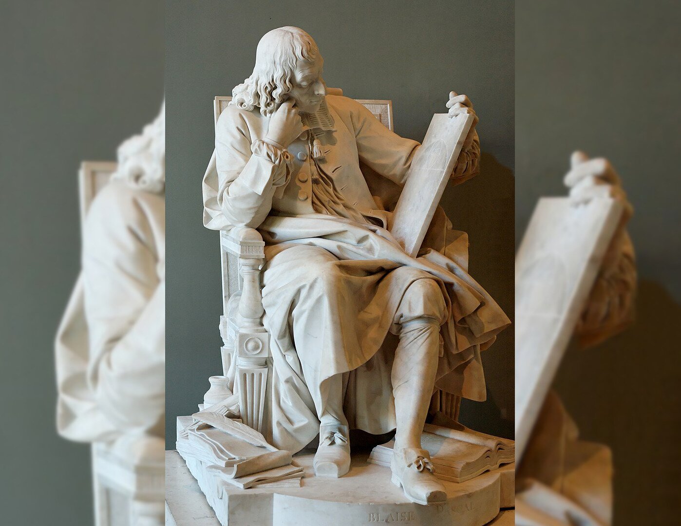 Zdjęcie przedstawia rzeźbę z białego marmuru, która prezentuje dojrzałego mężczyznę. Mężczyzna siedzi na krześle, ma długie, falujące włosy. Podpiera głowę ręką opartą o poręcz fotela. Ubrany jest w długi płaszcz z guzikami i szerokim kołnierzem. W ręku trzyma tablicę, na którą patrzy. Pod krzesłem leżą kartki oraz otwarta książka.