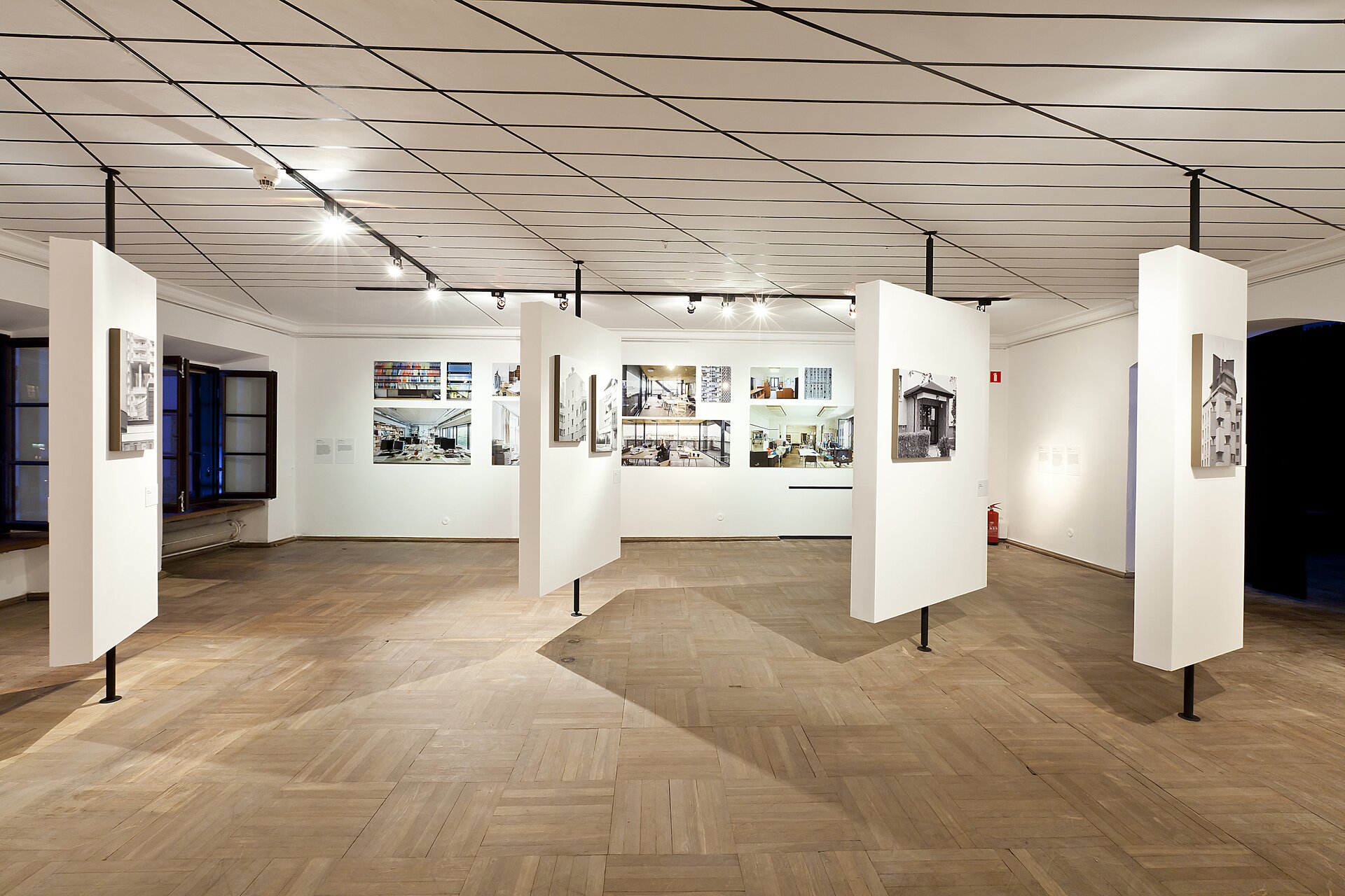 Na zdjęciu znajduje się pomieszczenie o biało‑czarnym suficie, brązowej podłodze i białych ścianach. Na suficie jest wiele poziomych i ukośnych czarnych linii oraz czarne szyny z małymi lampkami. Na ścianach wiszą kolorowe ilustracje o różnej wielkości. Z sufitu do podłogi poprowadzone są 4 rurki. Na każdej z nich zamontowany jest biały prostopadłościan, na którym powieszone są biało‑czarne obrazy. Na ścianie po lewej stronie znajdują się czarne okna ze szprosami. 