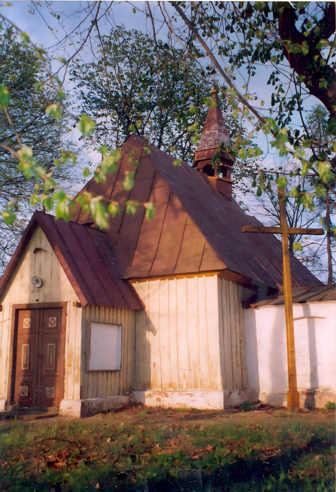 Zdjęcie przedstawia mały, drewniany kościół z brązowym, skośnym dachem. Na jego czubku znajduje się wieżyczka zakończona krzyżem. Przy kościele stoi drewniany krzyż. Ziemia pokryta jest zieloną trawą. W tle widoczne są drzewa z zielonymi liśćmi. Niebo jest błękitne. Zdjęcie zostało wykonane w świetle dziennym.