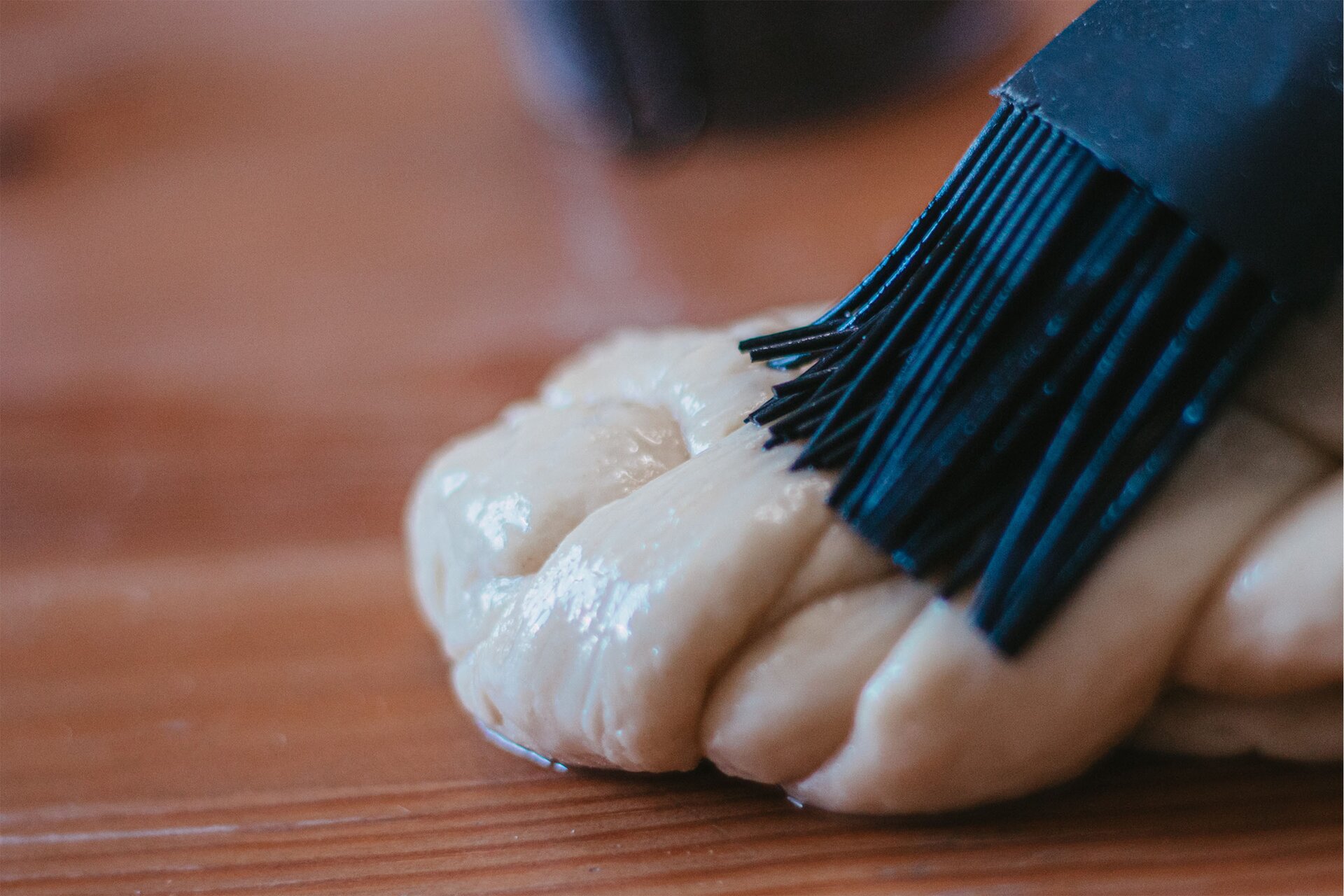 Zdjęcie przedstawia krok szósty formowania chały kijowskiej. Ujęcie wykonane jest od góry pod skosem. Widoczne jest zbliżenie na jeden z końców chały, która smarowana jest przezroczystym białkiem za pomocą silikonowego czarnego pędzelka kuchennego. Chałka znajduje się na ciemnobrązowym drewnianym blacie.