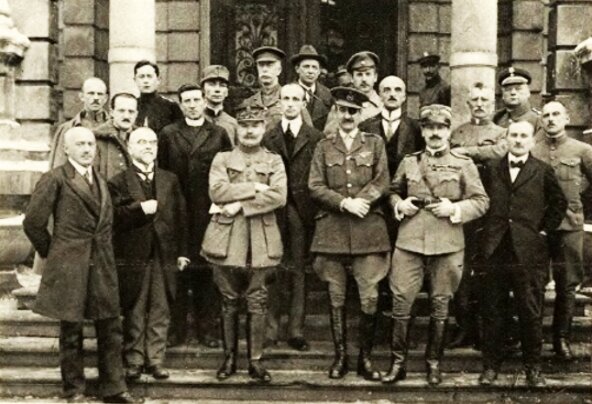 Fotografia przedstawia grupę mężczyzn stojących na schodach jednego z budynków. Niektórzy są ubrani w eleganckie ubrania, płaszcze i garnitury, inni są ubrani w mundury wojskowe. 