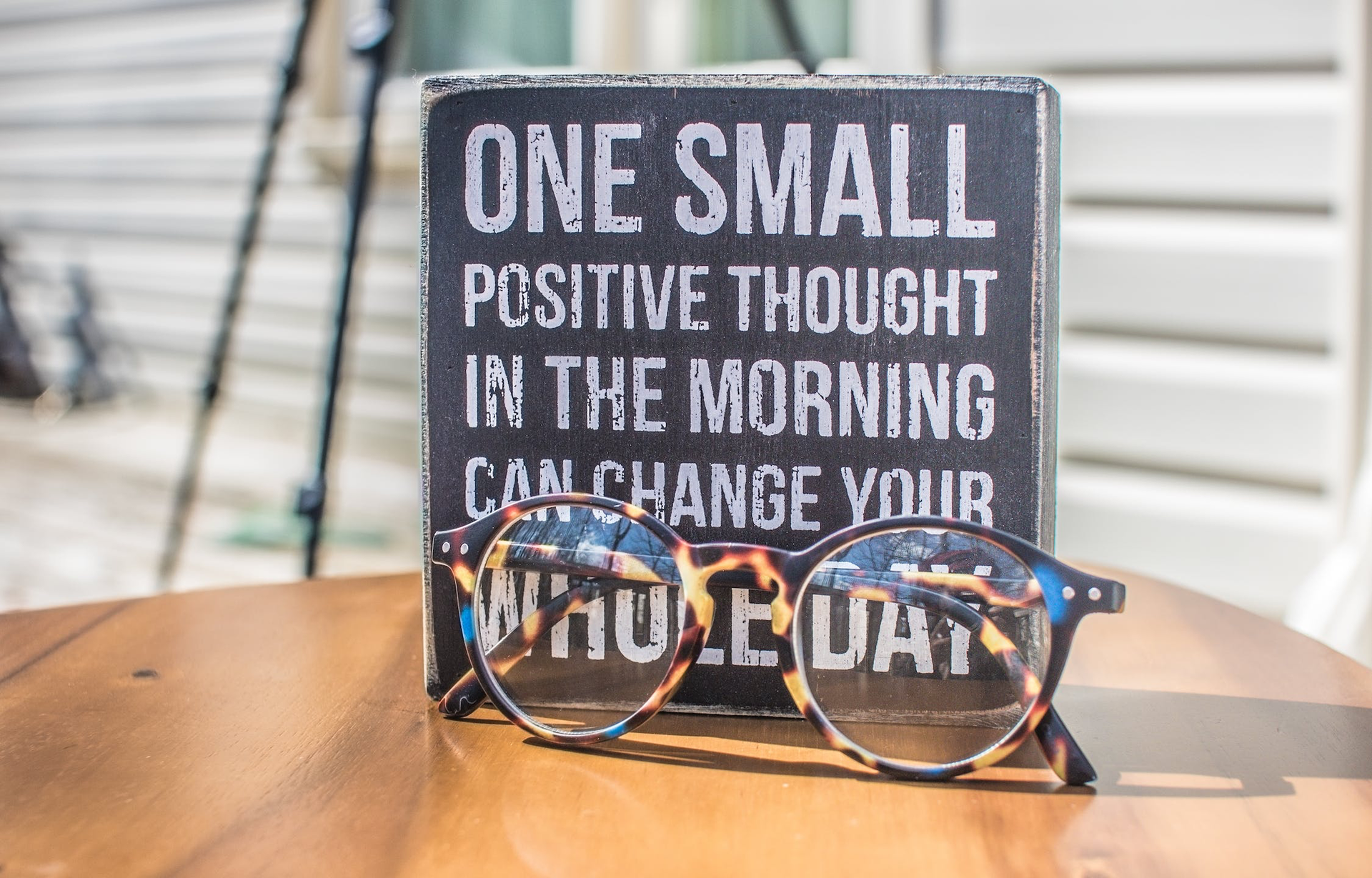 Zdjęcie przedstawia kwadratową tabliczkę stojącą na drewnianym blacie, przed którą widoczne są okulary korekcyjne. Na szarym tle tabliczki widoczny jest napis w języku angielskim: ONE SMALL POSITIVE THOUGHT IN THE MORNING CAN CHANGE YOUR WHOLE DAY.