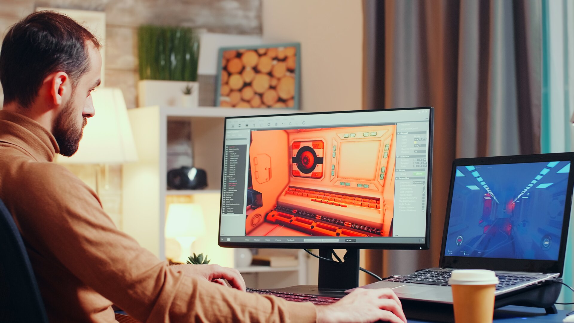 Fotografia przedstawia mężczyznę siedzącego przy biurku, na którym stoją dwa komputery stacjonarny i przenośny. Oba ekrany są włączone.