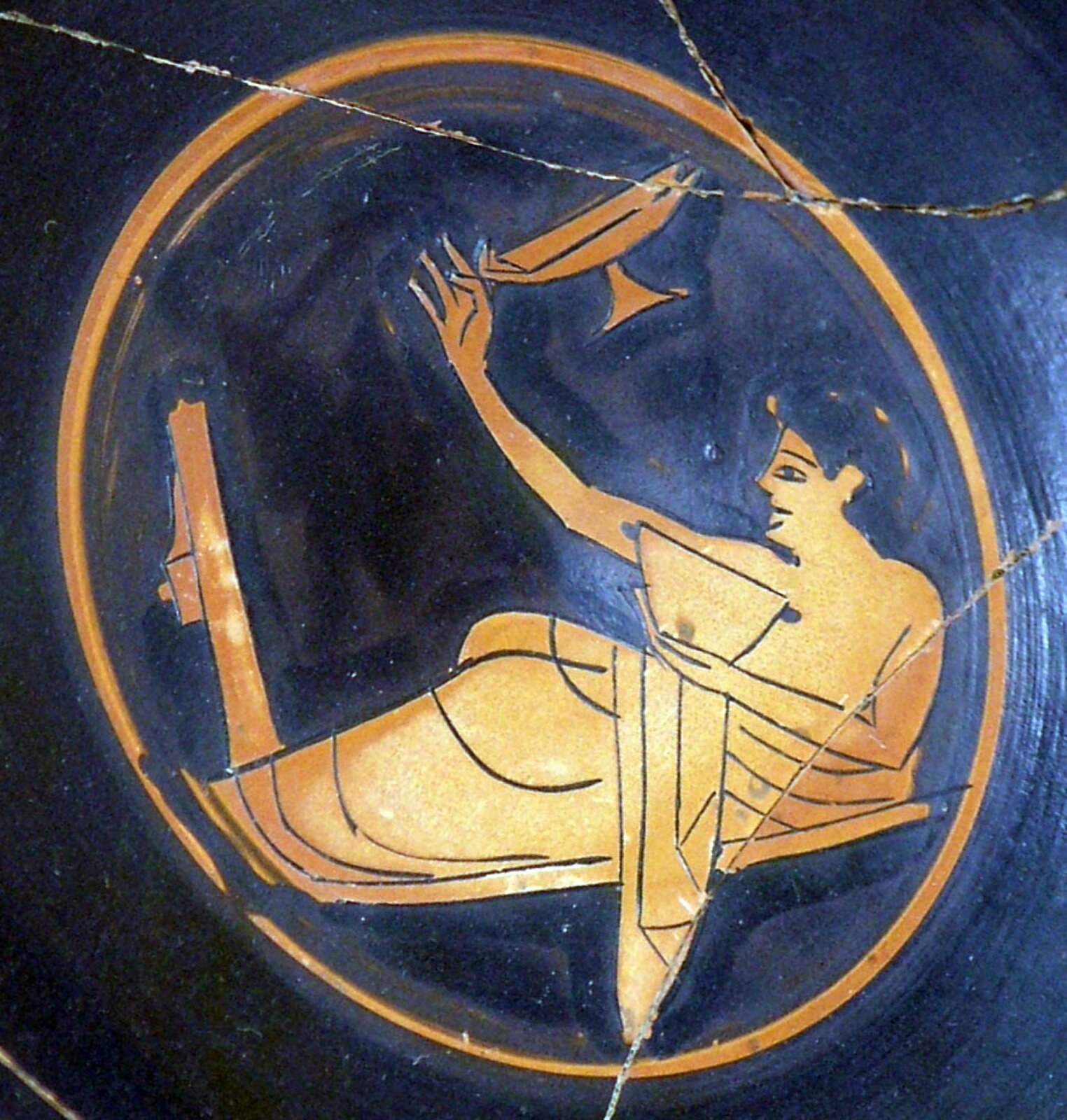 Ilustracja przedstawia młodego mężczyznę grającego w kottabos. Jest to scena z greckiego naczynia  z okresu ok. 510 r. p.n.e. Na granatowym zdjęciu w złotym okręgu został przedstawiony leżący, namalowany na złoto mężczyzna, który jest przedstawiony z lewego profilu. Prawą rękę ma podniesioną do góry, a tuż obok niej znajduje się złoty przedmiot w kształcie misy z nóżką. W lewej dłoni przy klatce piersiowej trzyma  złotą misę. Od pasa w dół jest odziany w szatę. Mężczyzna leży na wąskiej, poziomej powierzchni, a przy jego stopach znajduje się długi, pionowy pręt. 