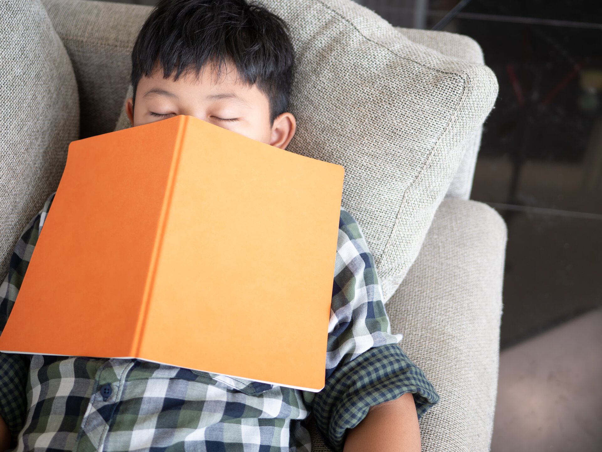 Zdjęcie przedstawia chłopca leżącego na szarej kanapie. Ma zamknięte oczy i położoną na twarzy książkę z pomarańczową okładką.