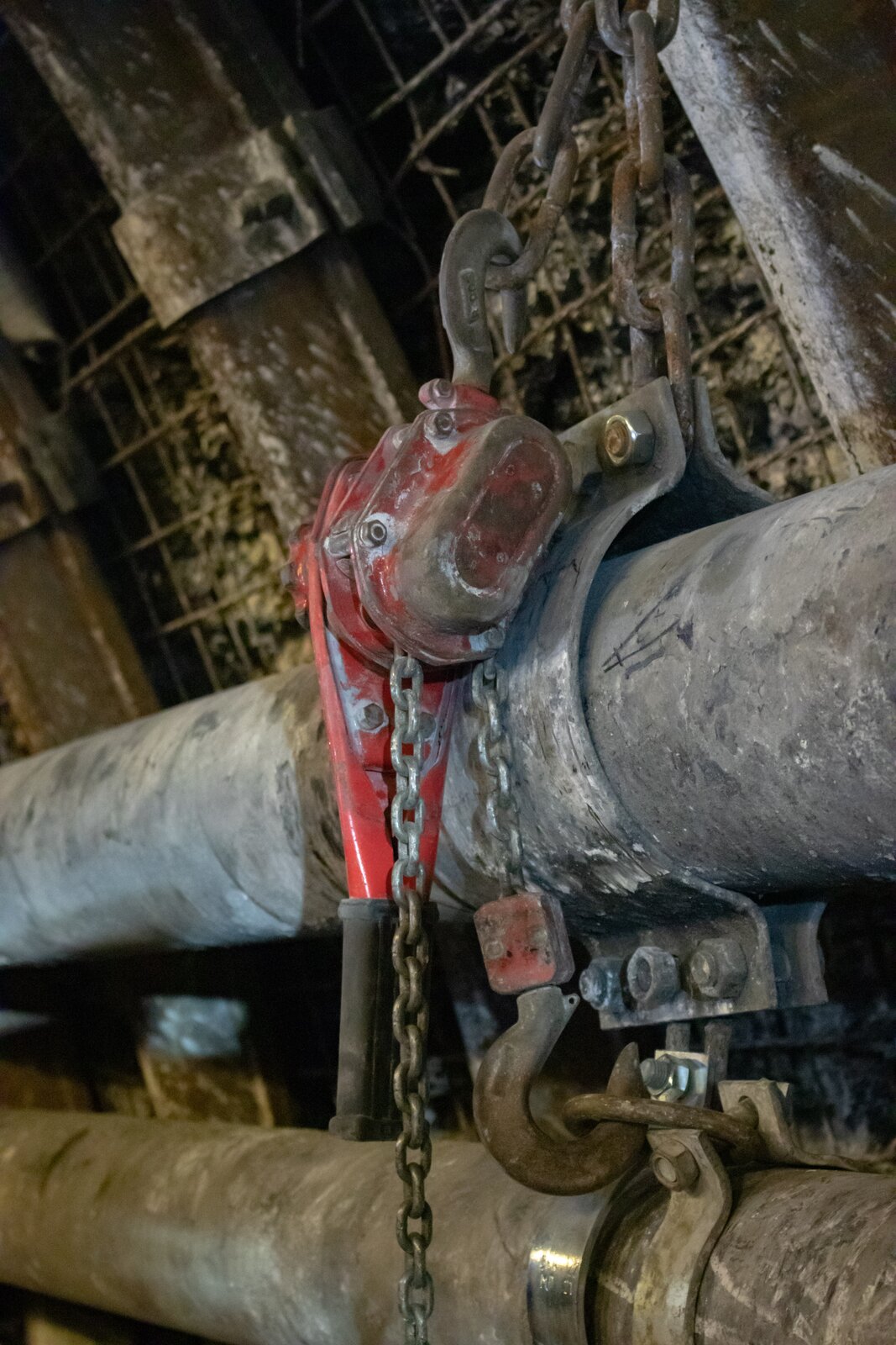 Na zdjęciu widać wciągarkę łańcuchową. Centralnym elementem zdjęcia jest stalowa rura. Rura jest między dwoma obejmami. Połączone obejmy są zawieszone na łańcuchu oczkowym. Łańcuch łączy się z dwoma eliptycznymi czerwonymi elementami połączonymi śrubami ze sobą. Pomiędzy elementami opadają dwa łańcuchy. Łańcuch po lewej stronie opada swobodnie. Łańcuch po prawej jest zaczepiony o stalowe ucho przy obejmie drugiej rury.