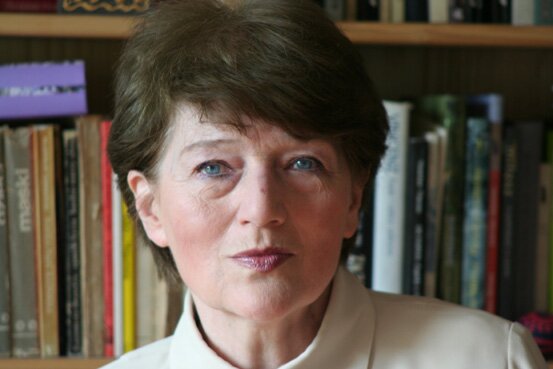 Na zdjęciu znajduje się kobieta na tle książek. Ma ciemne krótkie włosy, niebieskie oczy i poważny wyraz twarzy. Jest ubrana w jasną koszulę.