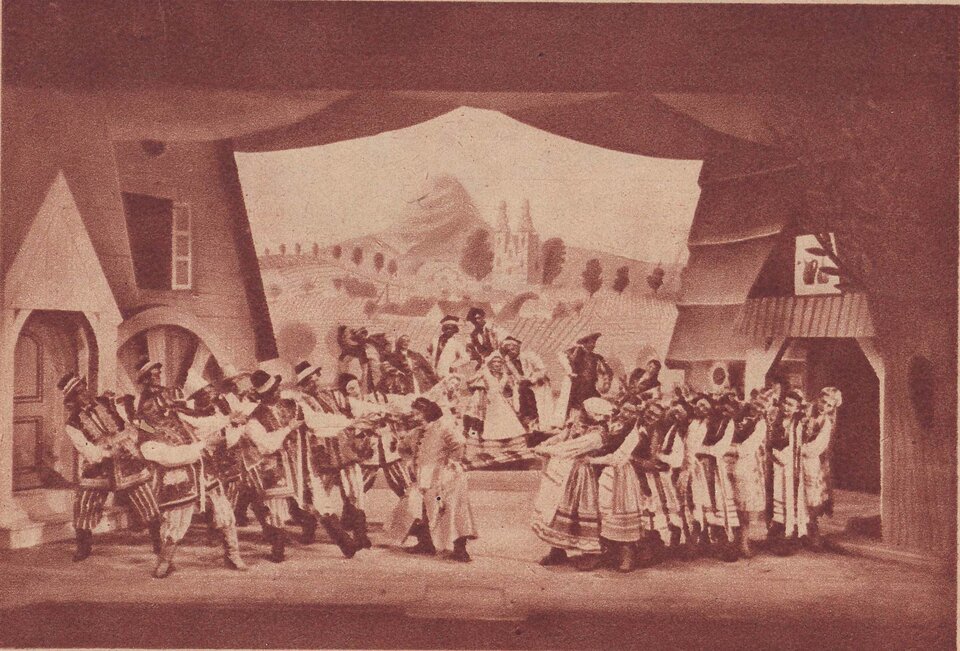 Fotografia przedstawia scenę teatralną. Tańczą tu liczne postaci ubrane w góralskie stroje. Scenografię stanowią fronty góralskich domów. W tle umieszczono planszę ukazującą górski pejzaż. 