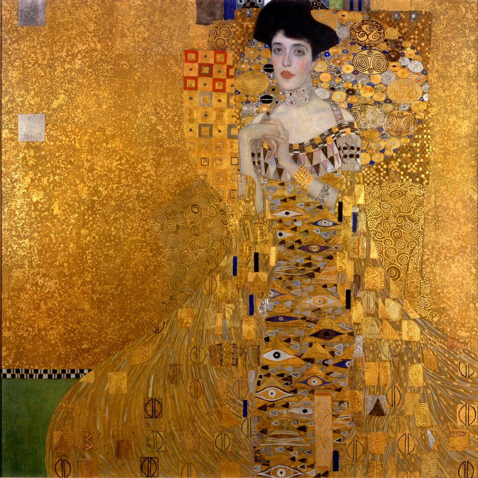 Obraz Gustava Klimta „Portret Adele Bloch‑Bauer I” przedstawia siedzącą kobietę na tle bogatej, złotej ornamentyki. Ma ciemne, wysoko upięte włosy, jasną twarz i usta podkreślone czerwoną barwą. Jej szyję zdobi szeroka, wysadzana kamieniami kolia, a na ramionach i przedramionach widoczne są bransolety. Kobieta ma na sobie długą, bogato zdobioną suknię, której wzór tworzą geometryczne figury, spirale, okręgi i stylizowane oczy w odcieniach złota, srebra, bieli, czerni i błękitu. Za jej głową znajduje się dekoracyjne, mozaikowe tło z licznymi spiralnymi i okrągłymi motywami. Lewą część obrazu wypełnia złote tło z geometrycznymi kwadratami. Dominującym kolorem kompozycji jest złoty.