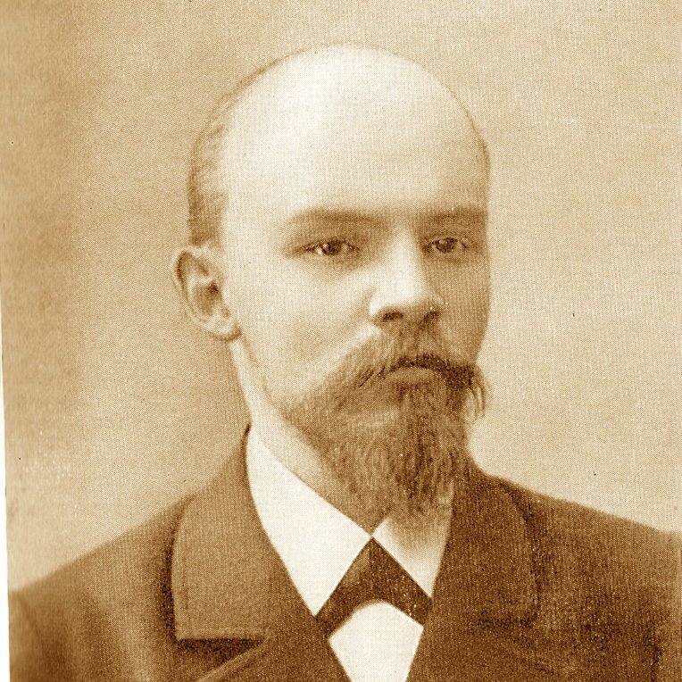 Fotografia przedstawia młodego mężczyznę pozującego do zdjęcia. Ma szczupłą twarz, małe oczy, jasne włosy z tyłu głowy i nad uszami, przód głowy jest łysy, jasne wąsy i krótką brodę. Ubrany jest w marynarkę i białą koszulę.