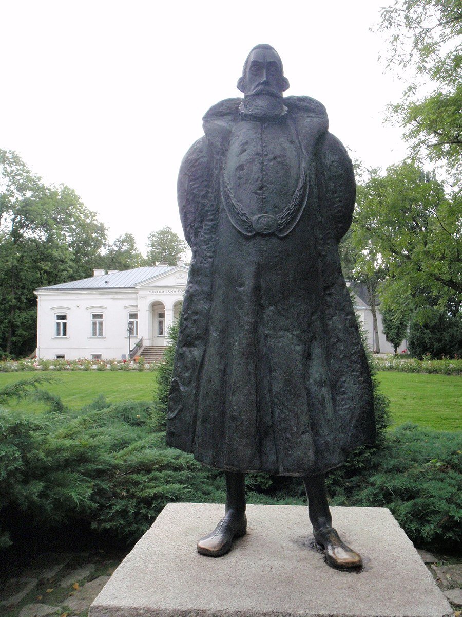 Zdjęcie przedstawia pomnik dojrzałego mężczyzny na tle parterowego dworku ustyuowanego wśród drzew. Mężczyzna ma pociągłą twarz, krótkie włosy oraz gęstą brodę z wąsami. Ubrany jest w długi żupan i płaszcz z szerokim kołnierzem, na szyi ma bogaty łańcuch. Mężczyzna trzyma ręce za plecami, jest wyprostowany. Pomnik stoi na betonowej podstawie, otaczają go niskie krzaki. 