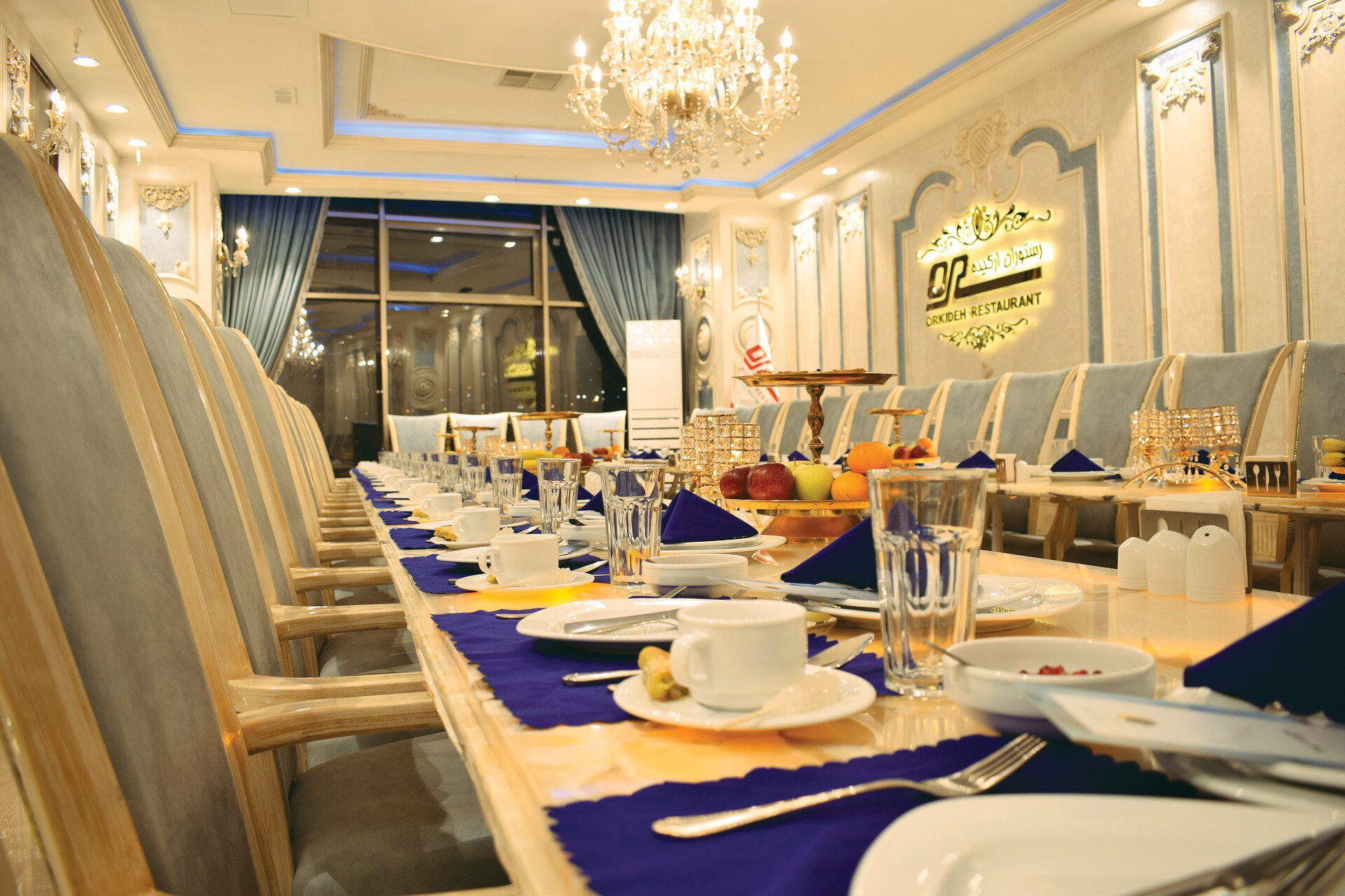 Zdjęcie przedstawia wnętrze eleganckiej sali restauracyjnej. Znajduje się w niej długi stół, na którym stoją nakrycia i sztućce. Przy stole stoją rzędami krzesła.