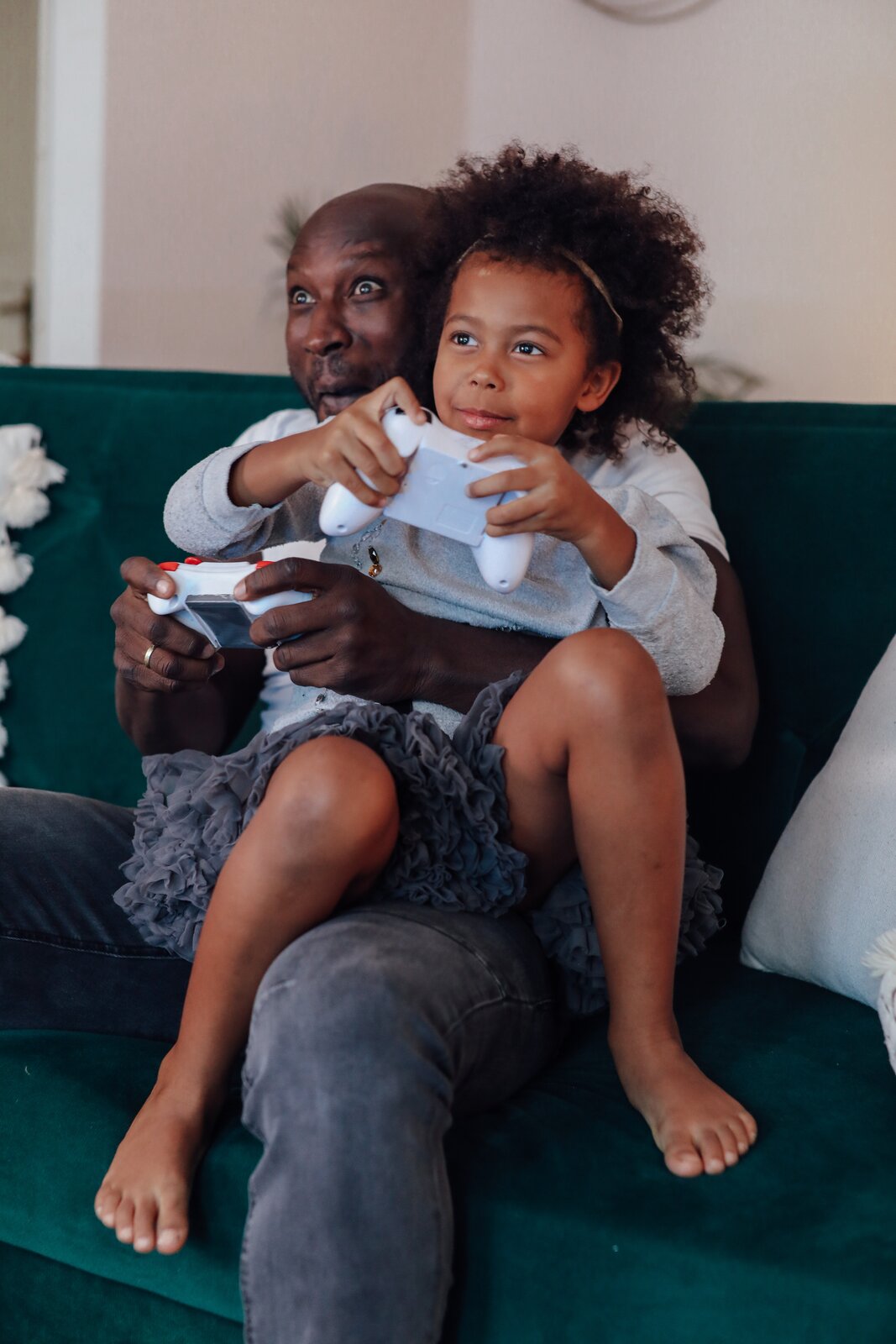 Zdjęcie przedstawia salon w domu. W tle widać beżową ścianę. Na pierwszym planie znajduje się ciemnozielona kanapa. Na kanapie siedzi ciemnoskóry, łysy mężczyzna ubrany w koszulkę z krótkimi rękawami i długie ciemnoszare spodnie. W rękach trzyma biały pad, czyli bezprzewodowe urządzenie służące do gry na konsoli. Na kolanach mężczyzny siedzi dziewczynka o kręconych ciemnobrązowych włosach. Jest ubrana w jasnoszarą bluzę z długimi rękawami i krótką ciemnoszarą spódniczkę z falbankami. W rękach trzyma drugi pad. Mężczyzna i dziewczynka patrzą na niewidoczny na zdjęciu telewizor i się uśmiechają.