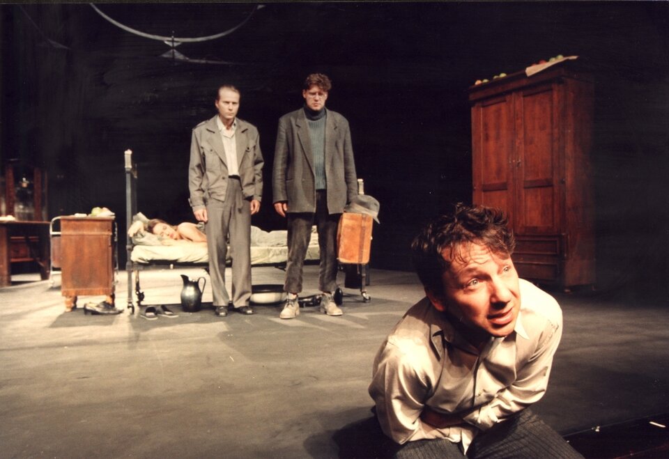 Zdjęcie ze spektaklu pt. "Kartoteka". Na pierwszym planie widać siedzącego, zgiętego w pół mężczyznę. Ubrany jest w białą koszulę i szare spodnie. Na środku sceny stoi metalowe łóżko, w którym śpi kobieta. Przed łóżkiem stoją dwaj mężczyźni. Mają zmęczone, smutne wyrazy twarzy. Jeden z nich ubrany jest w beżową kurtkę i beżowe spodnie. Drugi ma na sobie brązową marynarkę, brązowe spodnie i szary sweter. W ręku trzyma walizkę i kapelusz. Po prawej stronie pomieszczenia widać dużą szafę, po lewej- komodę.