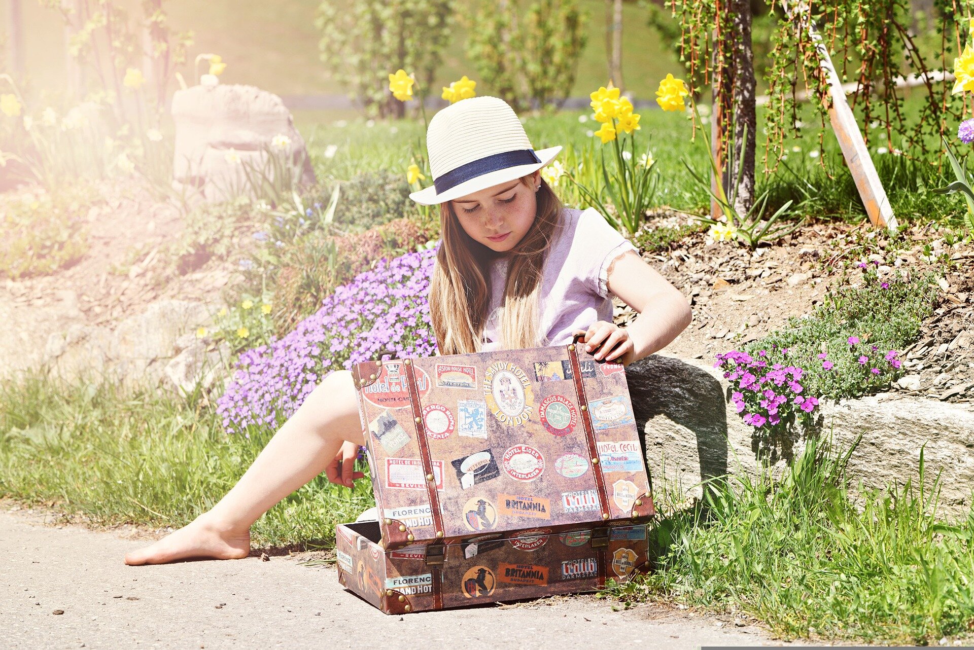 Zdjęcie przedstawia dziewczynę siedzącą z walizką w ogrodzie.