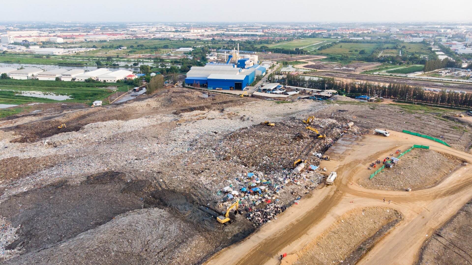 Ilustracja przedstawia składowisko odpadów znajdujące się poza miastem. Na skraju składowiska widoczny budynek. 