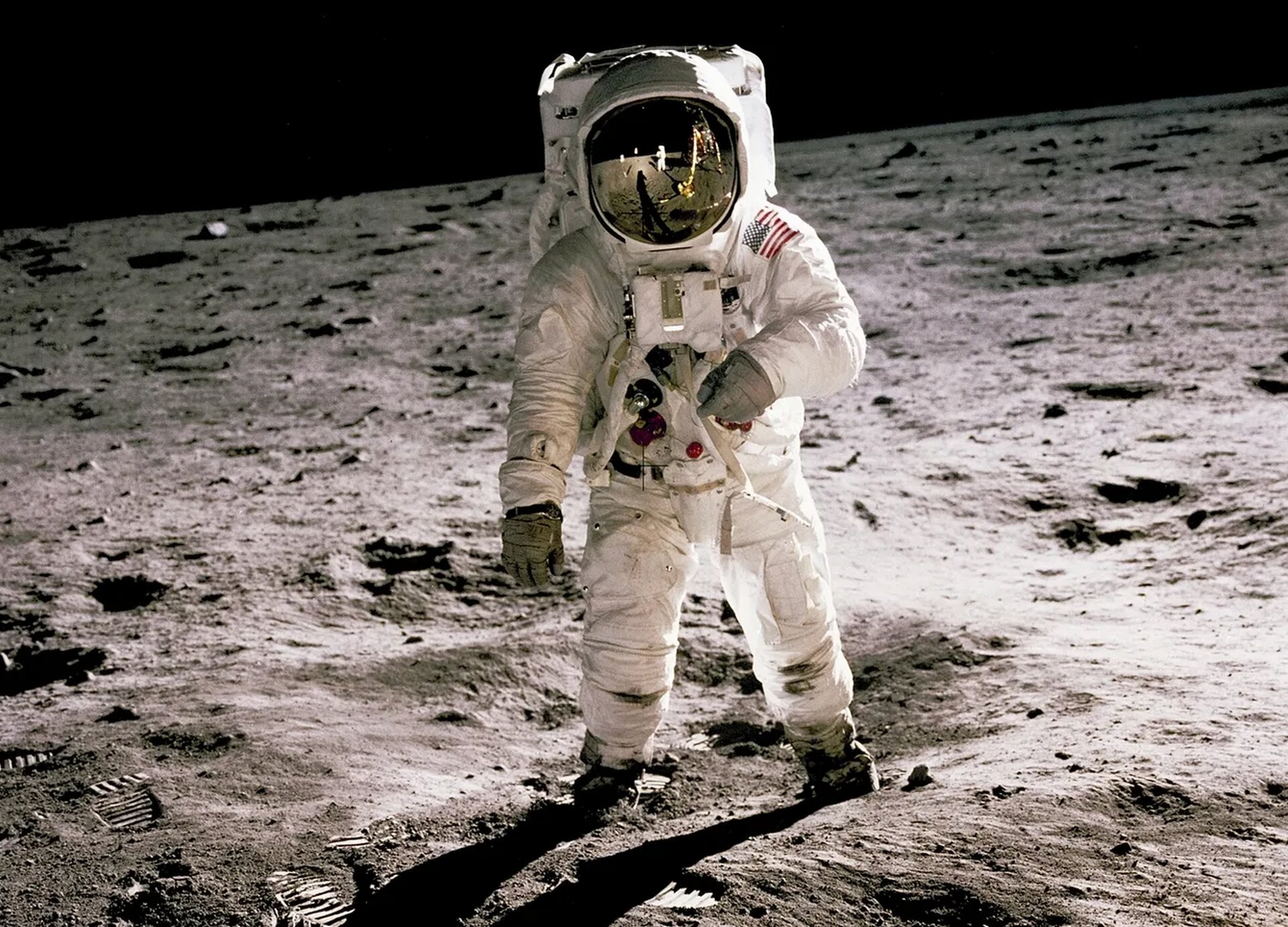  Zdjęcie przedstawia postać w białym skafandrze astronauty, z hełmem z zamkniętą szybką odbijającą światło oraz z plecakiem z tlenem na plecach. Na lewym ramieniu skafandra widoczna jest naszywka z flagą amerykańską. Osoba stoi w rozkroku,  na pylastej i szarej powierzchni księżyca.