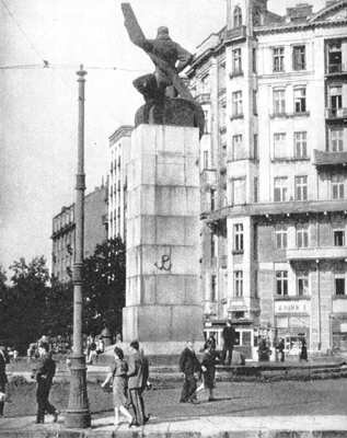 Czarno‑białe zdjęcie przedstawia pomnik znajdujący się na wysokim kamiennym cokole, na którym znajduje się namalowany symbol Polski Walczącej. Pomnik przedstawia mężczyznę trzymającego śmigło samolotu. Wokół pomnika spacerują ludzie. W oddali znajdują się kamienice, przed którymi rosną drzewa.