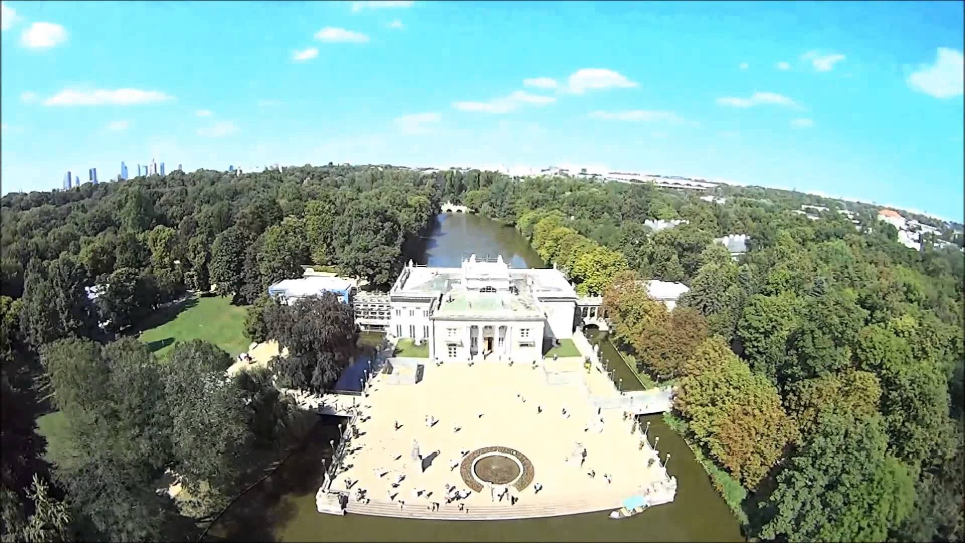 Ilustracja przedstawia widok z lotu ptaka na Łazienki Królewskie w Warszawie, tutaj widok na Pałac na Wodzie, który znajduje się w centralnym punkcie zdjęcia. Po bokach budynku widać tereny zielone. W oddali można zauważyć panoramę miasta.