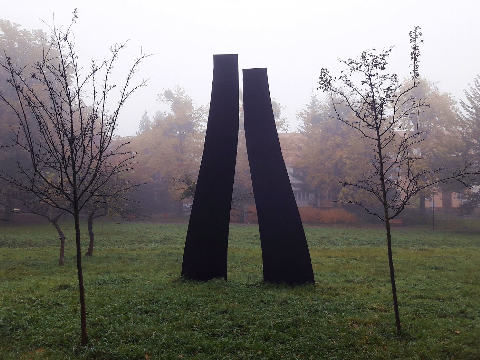 Na fotografii znajdują się 2 pionowe, podłużne, lekko wygięte formy w kolorze ciemnobrązowym. Dookoła jest trawnik i rosną pojedyncze, małe drzewa liściaste. W tle są wysokie drzewa z żółto‑brązowymi liśćmi i szary budynek. Niebo jest zachmurzone.