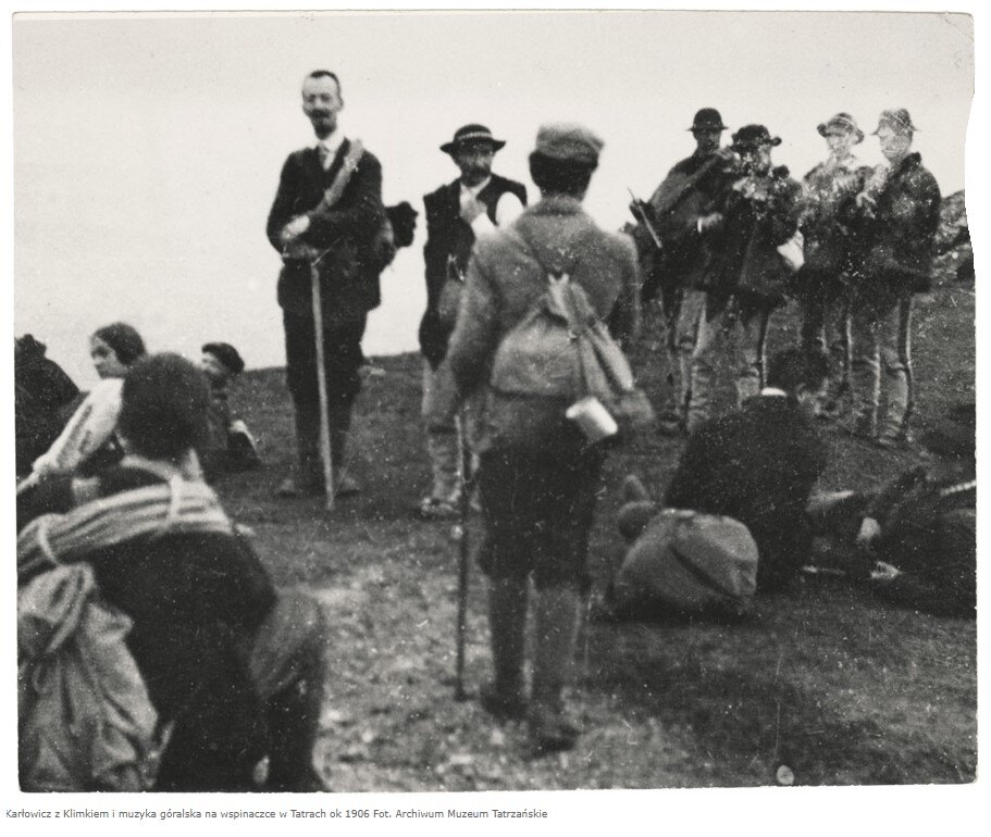 Stara fotografia przedstawia grupę ludzi podczas wędrówki górskiej, cześć z nich odpoczywa siedząc na ziemi, na drugim planie orkiestra góralska złożona z kilku mężczyzn w strojach górali podhalańskich.