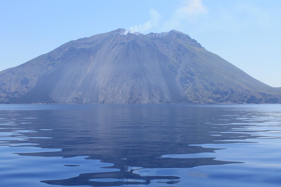 Na zdjęciu jest wulkan położony nad morzem. Na szczycie wulkanu widoczny jest krater, z którego unosi się cienka smuga dymu.  