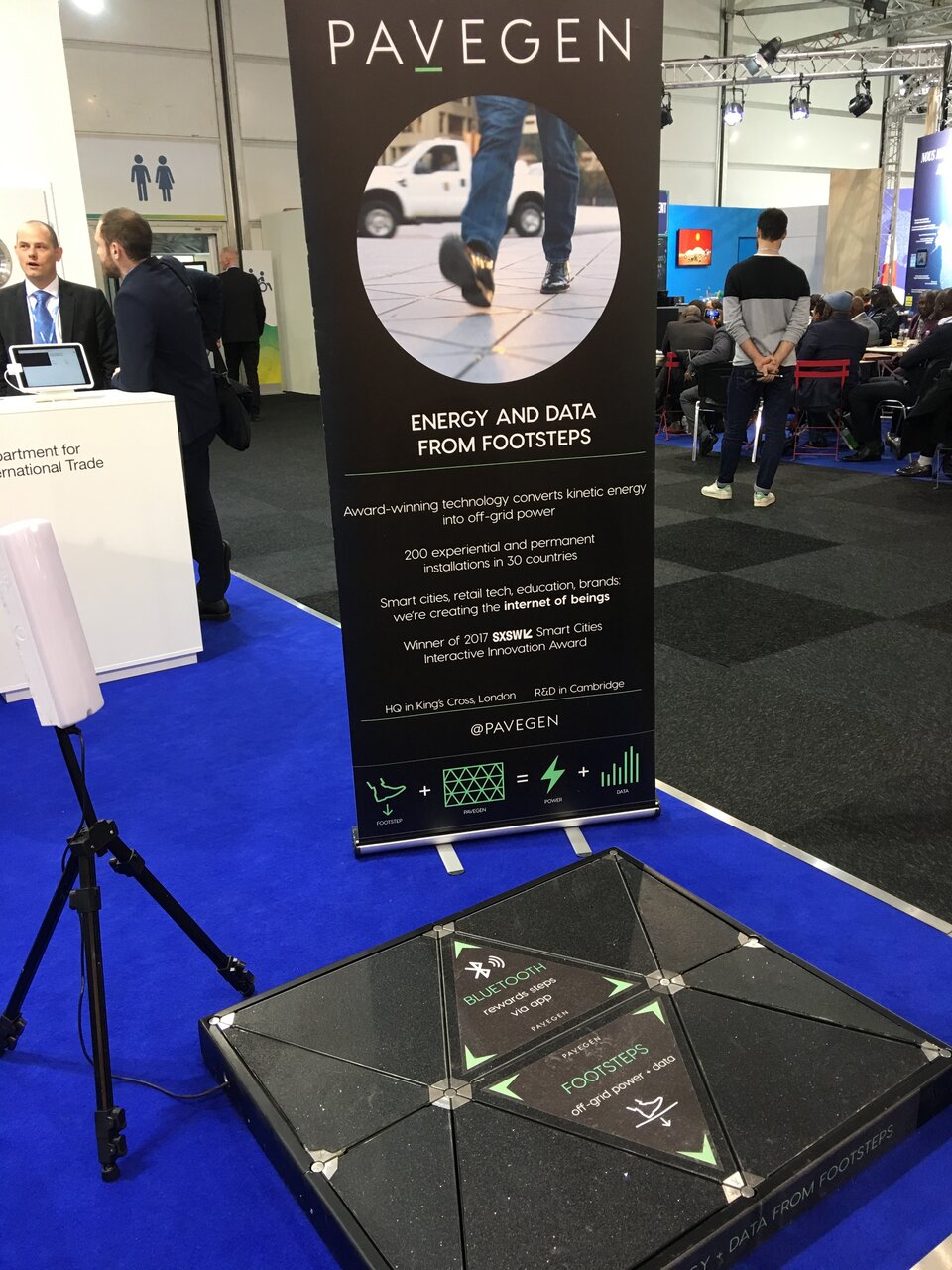 Zdjęcie przedstawia stoisko na targach, na którym przedstawione są koncepcyjne płyty chodnikowe w Londynie przekształcające energię kinetyczną ludzkich stóp w energię elektryczną (System PaveGen). W centrum znajduje się stojak z plakatem reklamowym z napisami i okrągłym zdjęciem  przedstawiającym płyty chodnikowe w kształcie trójkąta oraz widoczne nogi mężczyzny w jeansach i czarnych, skórzanych butach. Stojak znajduje się na niebieskiej wykładzinie, a przed nim widoczna jest instalacja ułożona z tych płyt. W tle czarna wykładzina i ludzie znajdujący się na innym stoisku. 