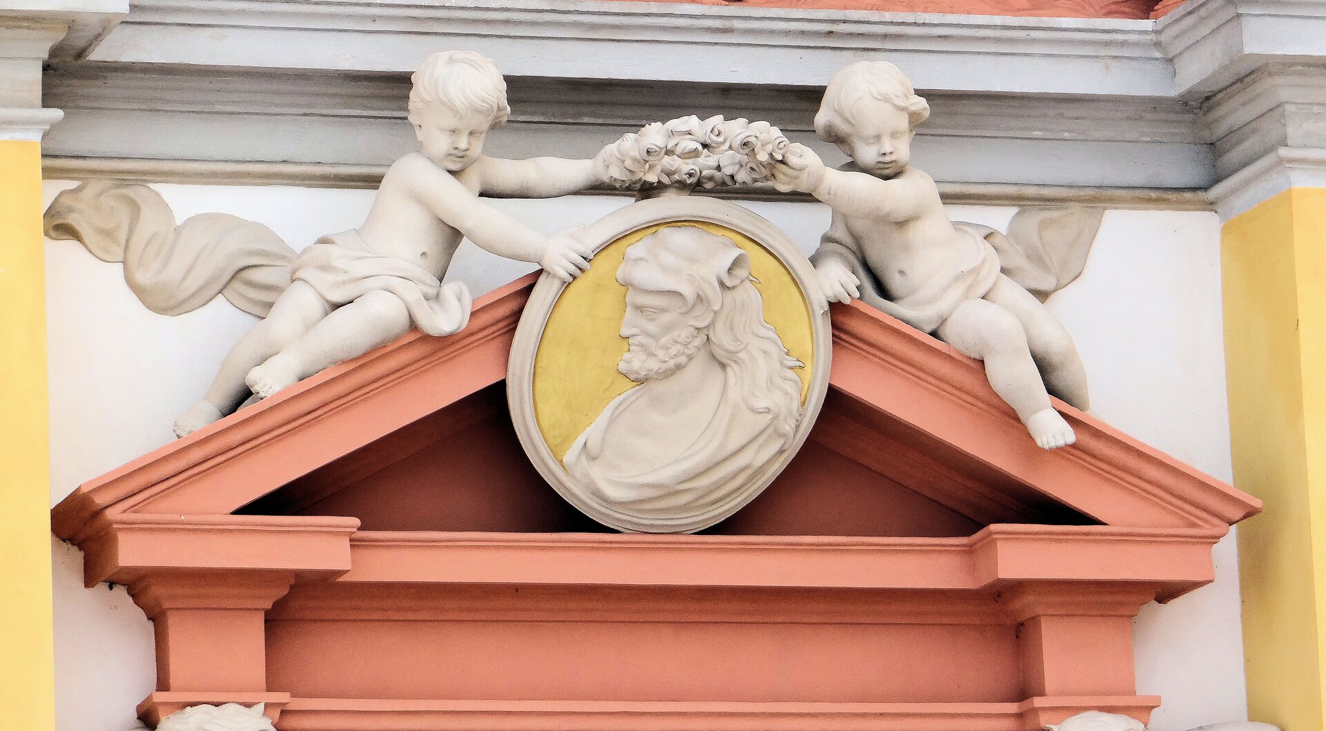 Fotografia Jolanty Dyr przedstawia widok na Medalion w tympanonie fasady pałacu Jana III Sobieskiego. Medalion przedstawia postać mężczyzny w średnim wieku, który ma długie włosy i bujny zarost. Podobizna mężczyzny znajduje się na białym tle, a sam medalion trzymany jest przez dwóch kupidynów – jednymi dłoniami trzymają medalion, a drugimi koronę. 
