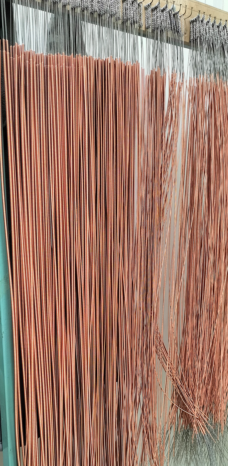 Zdjęcie przedstawia struny basowe wykonane z drutu miedzianego nawiniętego na rdzeń z drutu stalowego. Widoczne są struny zawieszone pękami na gwoździach wbitych w deskę.