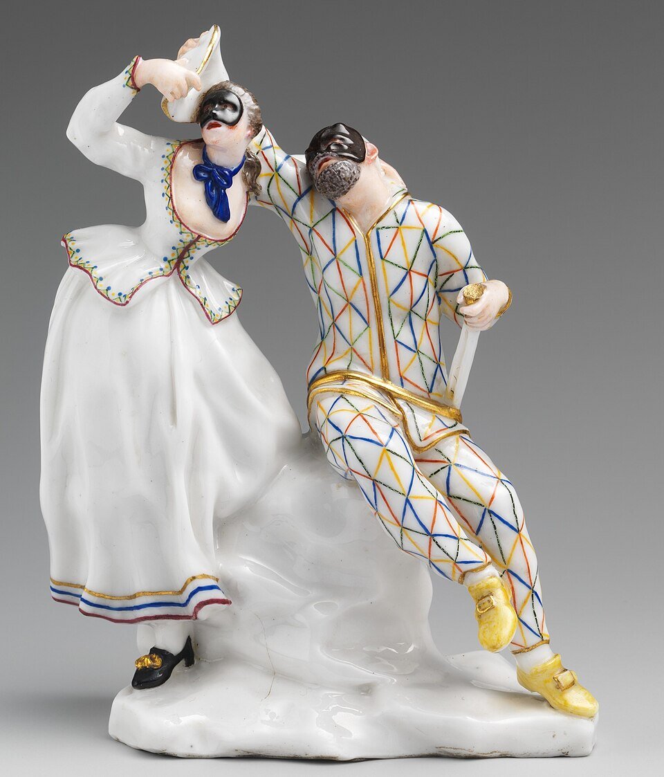 Ilustracja przedstawia figurkę z porcelany, której autorem jestGiuseppe Gricci. Figurka ukazuje dwie postacie w charakterystycznych strojach commedii dell’arte  – Harlekina i Kolombinę. Harlekin ubrany jest w kostium w romby w kolorach niebieskim, czerwonym i żółtym, z dodatkami w kolorze złotym. Na twarzy ma czarną maskę, a jego postawa jest swobodna, oparta o siedzenie. Kolombina ubrana jest w długą, białą suknię z kolorowymi lamówkami i niebieską kokardą pod szyją. Na głowie ma czapeczkę, a na twarzy czarną maskę. Jej uniesiona ręka sugeruje element teatralnej gry lub flirtu.  Figurka wykonana jest z porcelany, starannie malowanej ręcznie. Złocenia i precyzyjne detale podkreślają wysoką jakość rzemiosła. Całość osadzona jest na białej, falistej podstawie.	